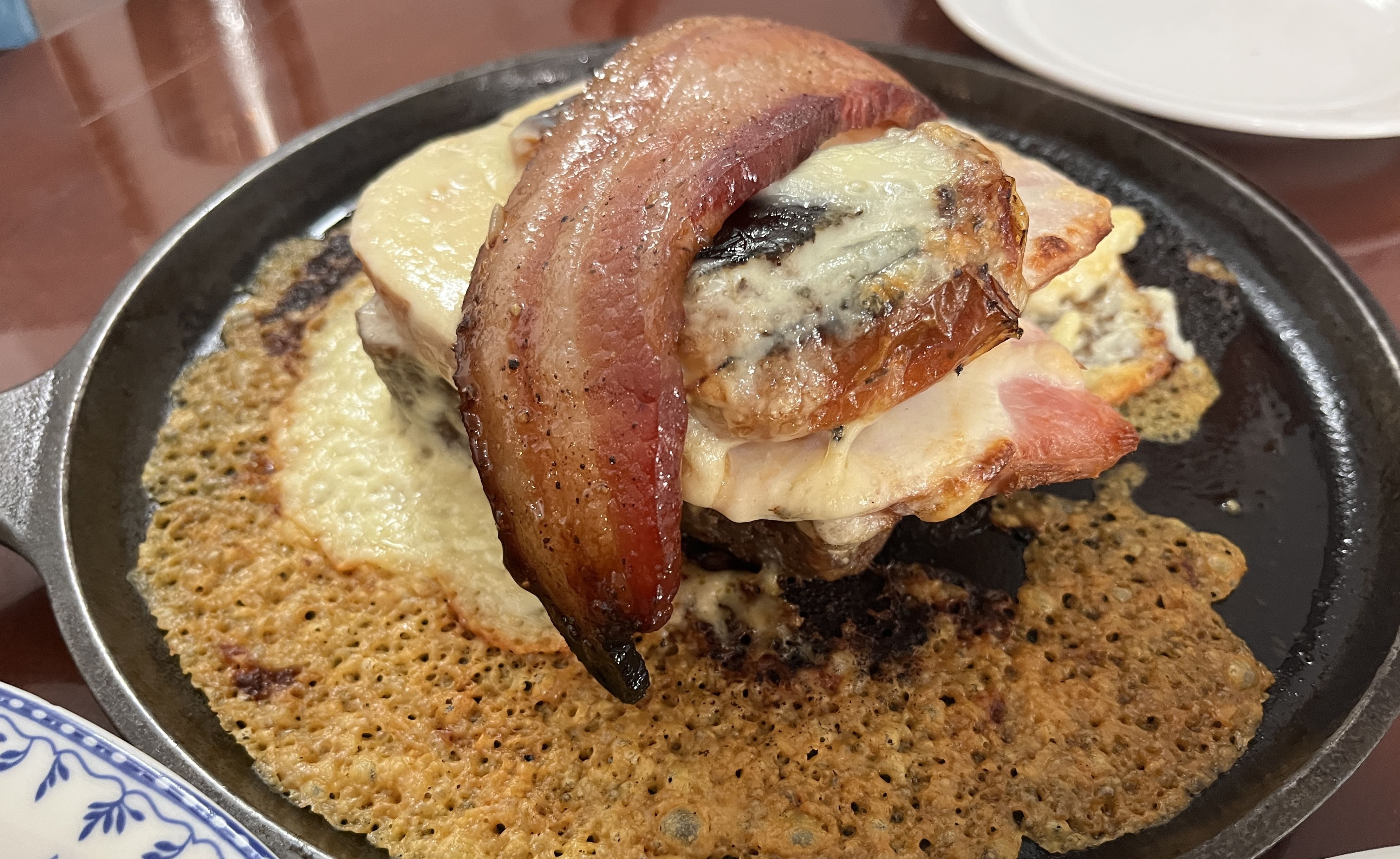 Close-up of a sizzling cast iron skillet with melted cheese, crispy bacon, grilled mushrooms, and a thick slice of ham on top, against a reddish-brown table surface.