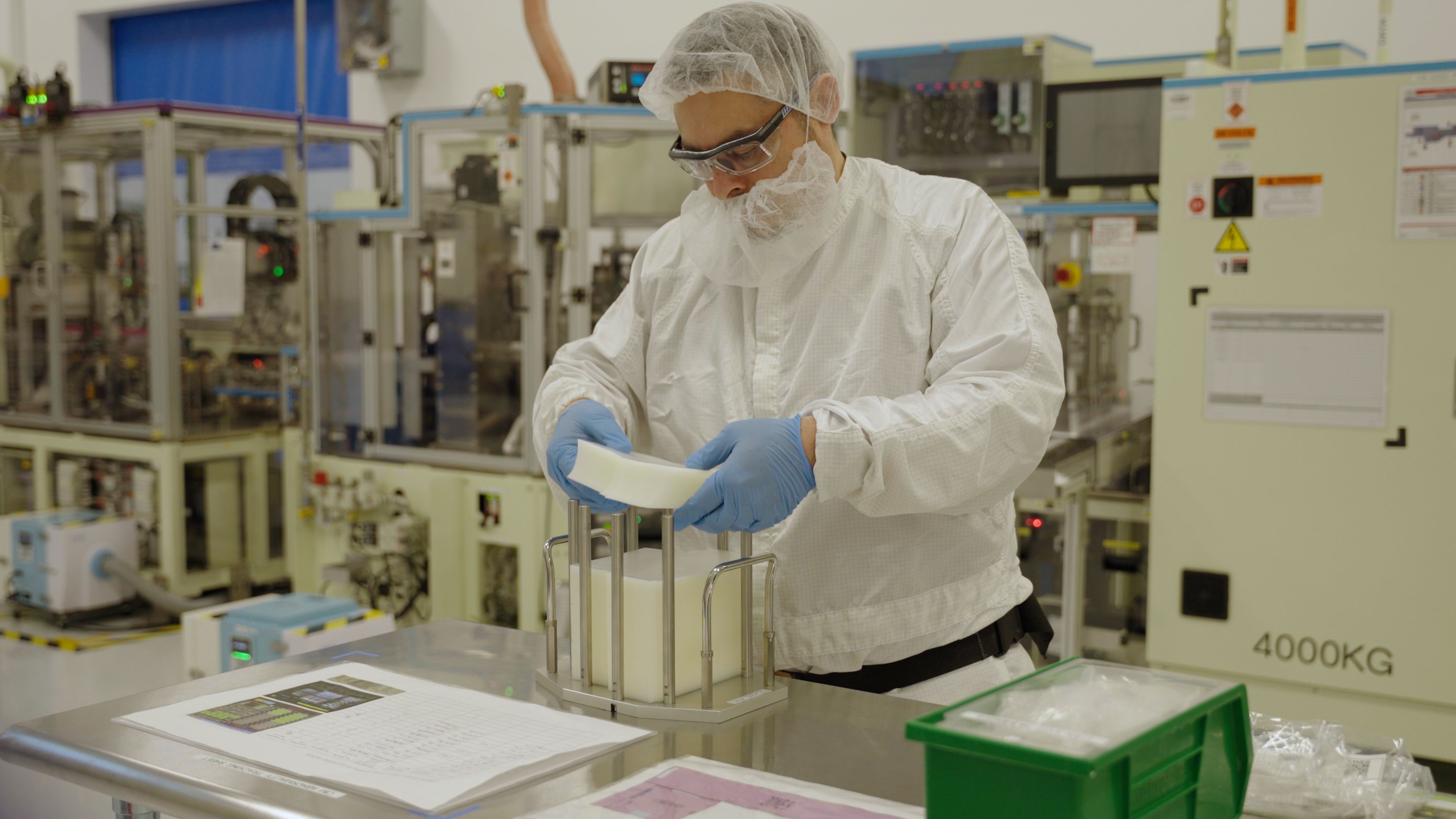 A worker in white protective gear, gloves, hairnet, and goggles inspects white foam sheets in a clean industrial setting with machinery in the background.