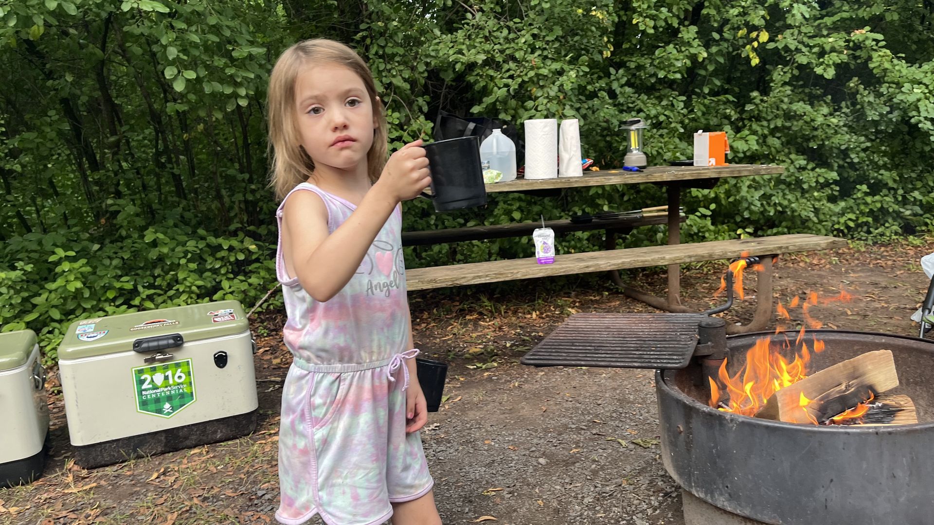 A girl holds a cup while standing next to a campfire