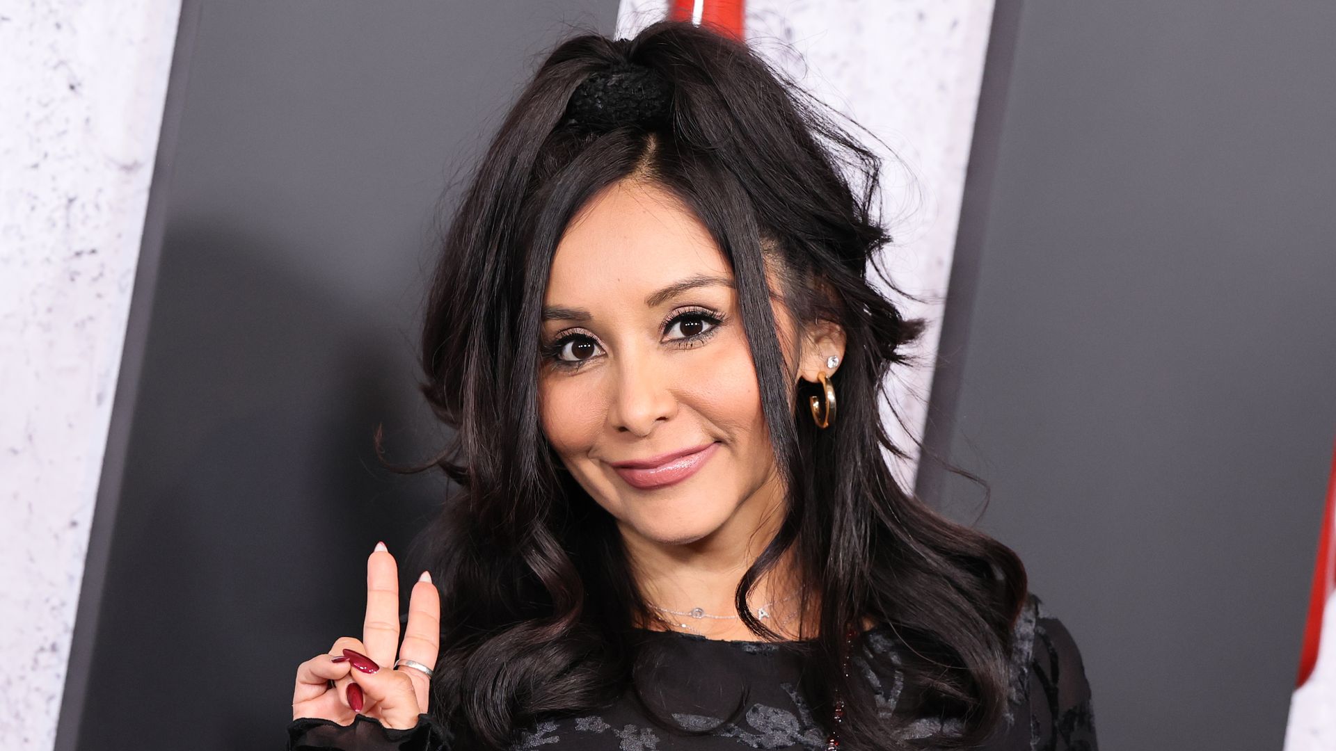 Smiling woman with long dark hair in a high ponytail, wearing a black patterned dress, gold hoop earring, and pink lipstick; she shows a peace sign with red nails against a gray backdrop.