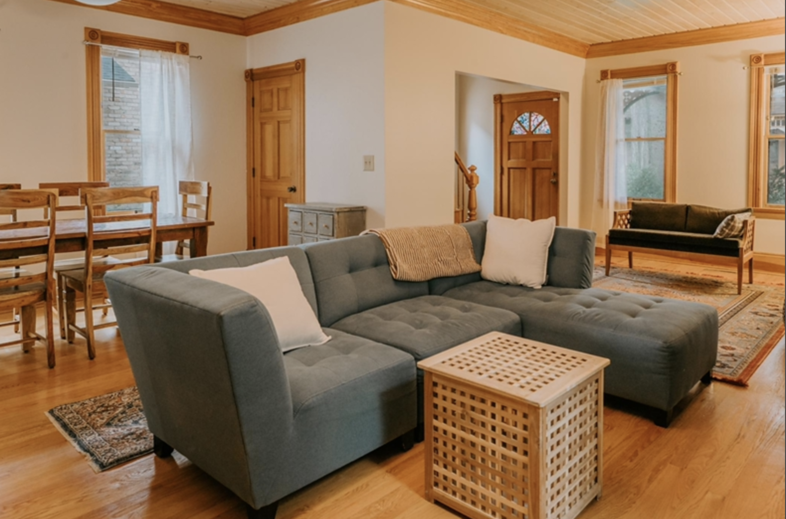 Cozy living room with gray sectional sofa, white pillows, beige throw, wooden coffee table, dining table with chairs, wooden floors and doors, soft natural light from windows.