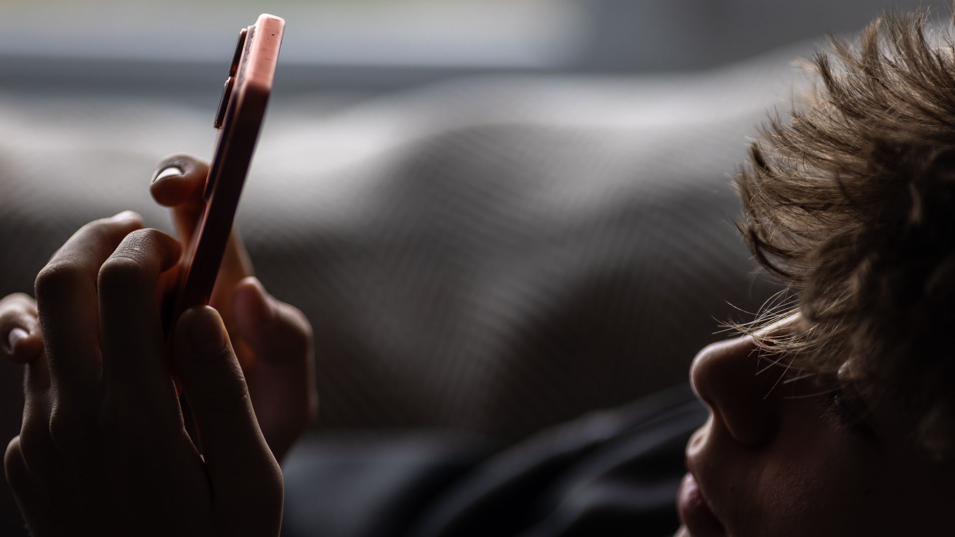 Close-up of a person holding a pink smartphone with both hands in a dim room, while a child with messy brown hair sits nearby, blurred in the background.