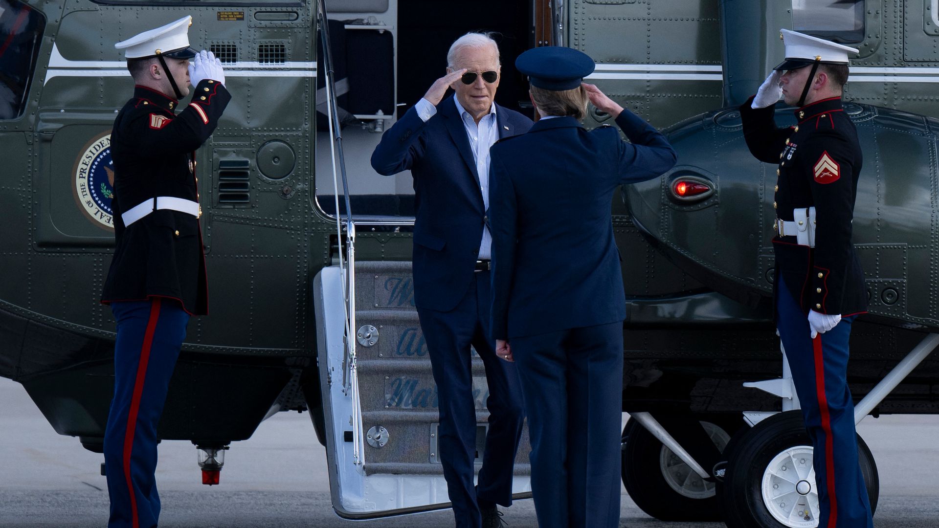 President Biden, wearing a blue suit, salutes a soldier while flanked by two more soldiers while stepping out of his green Marine One helicopter.