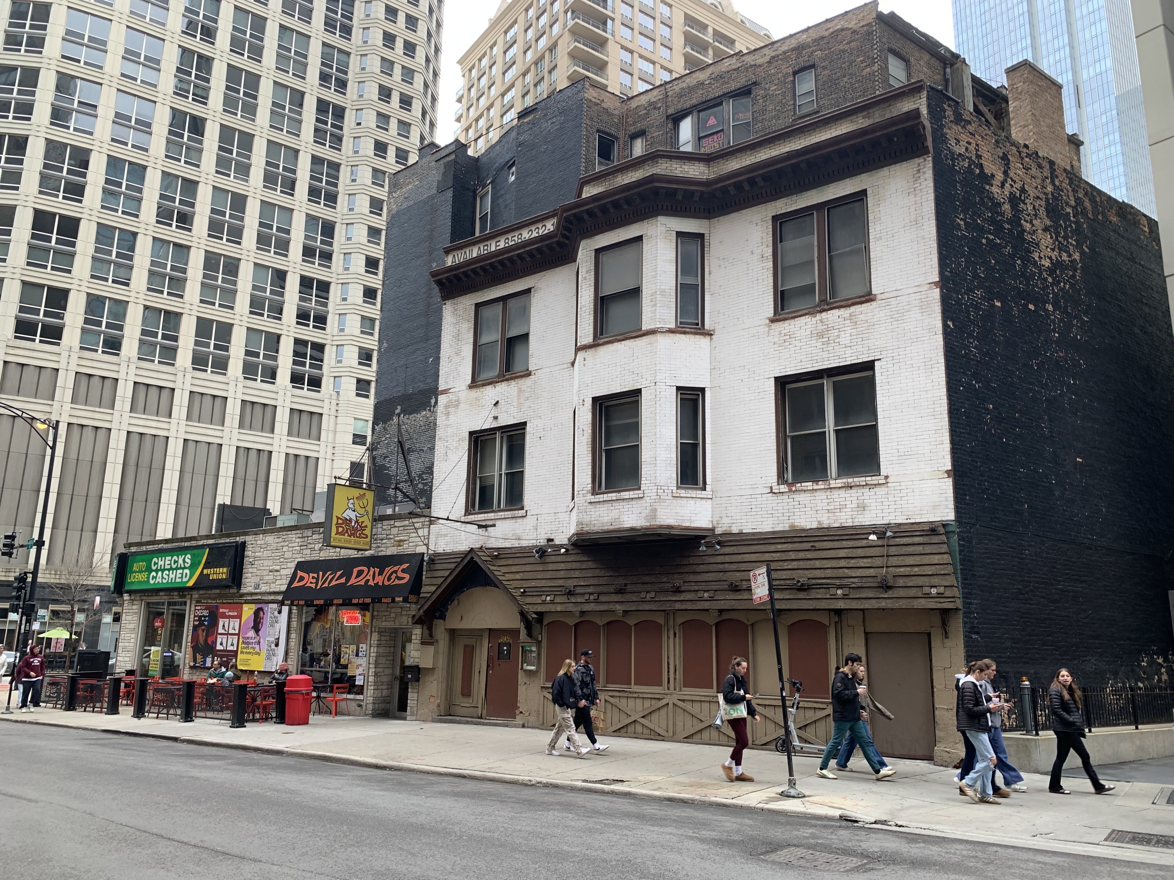 Urban street scene with old three-story white brick building, adjacent businesses including "Devil Dawgs" hot dog shop, and several pedestrians walking on the sidewalk.