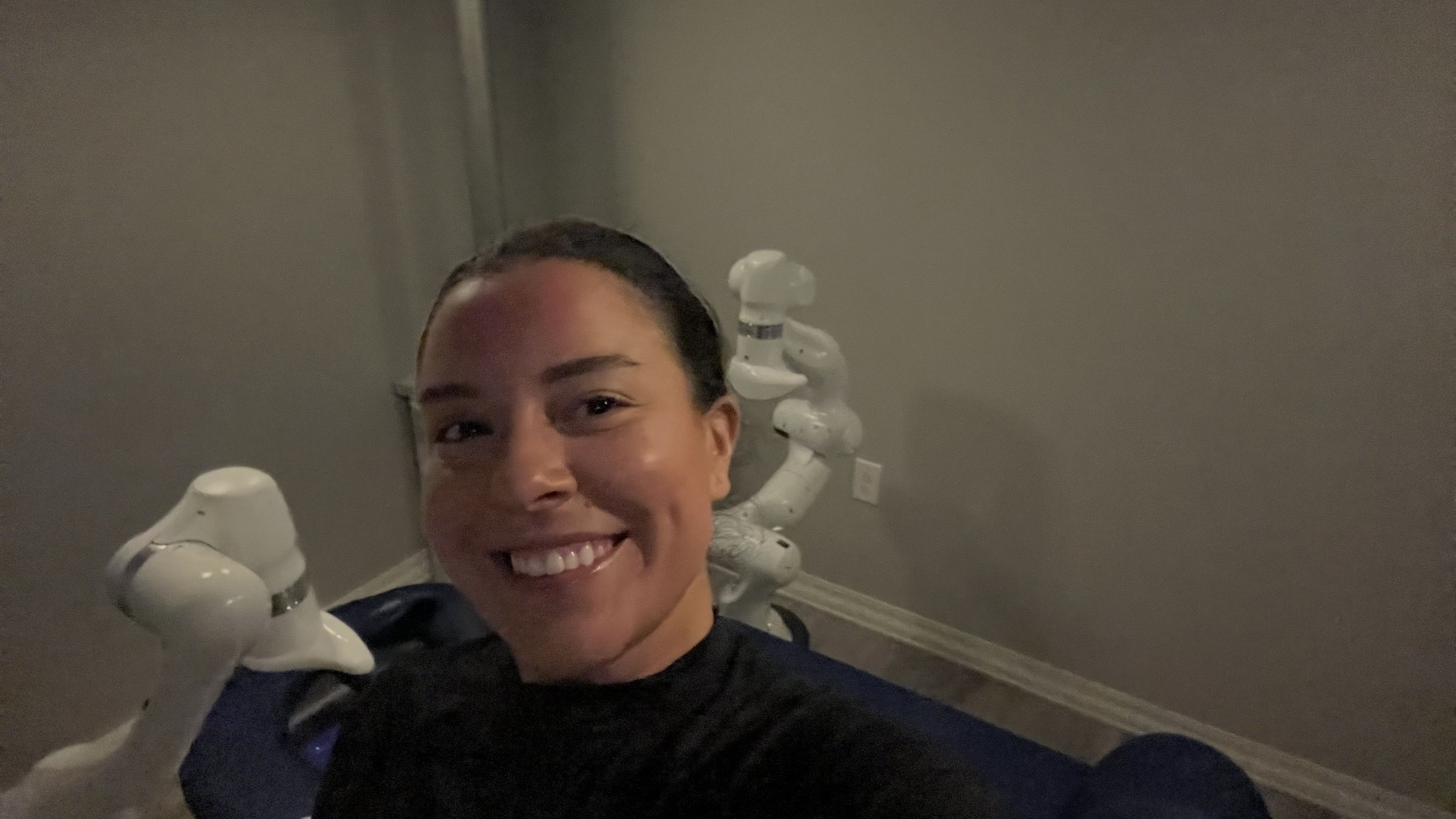 Smiling woman with dark hair wearing a black top, taking a selfie indoors next to a white robotic arm against a plain gray wall.