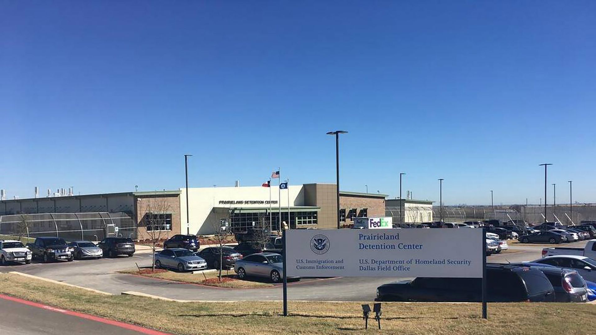 Exterior view of Prairieland Detention Center under clear blue sky, with parking lot full of cars and a sign showing U.S. Immigration and Customs Enforcement and Homeland Security logos.