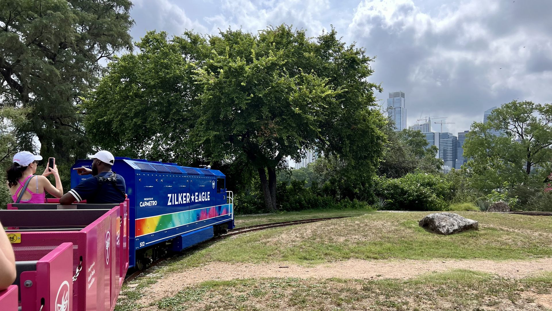 Two people sit at the front of the train, which reads "Zilker Eagle" with the skyline in the background.