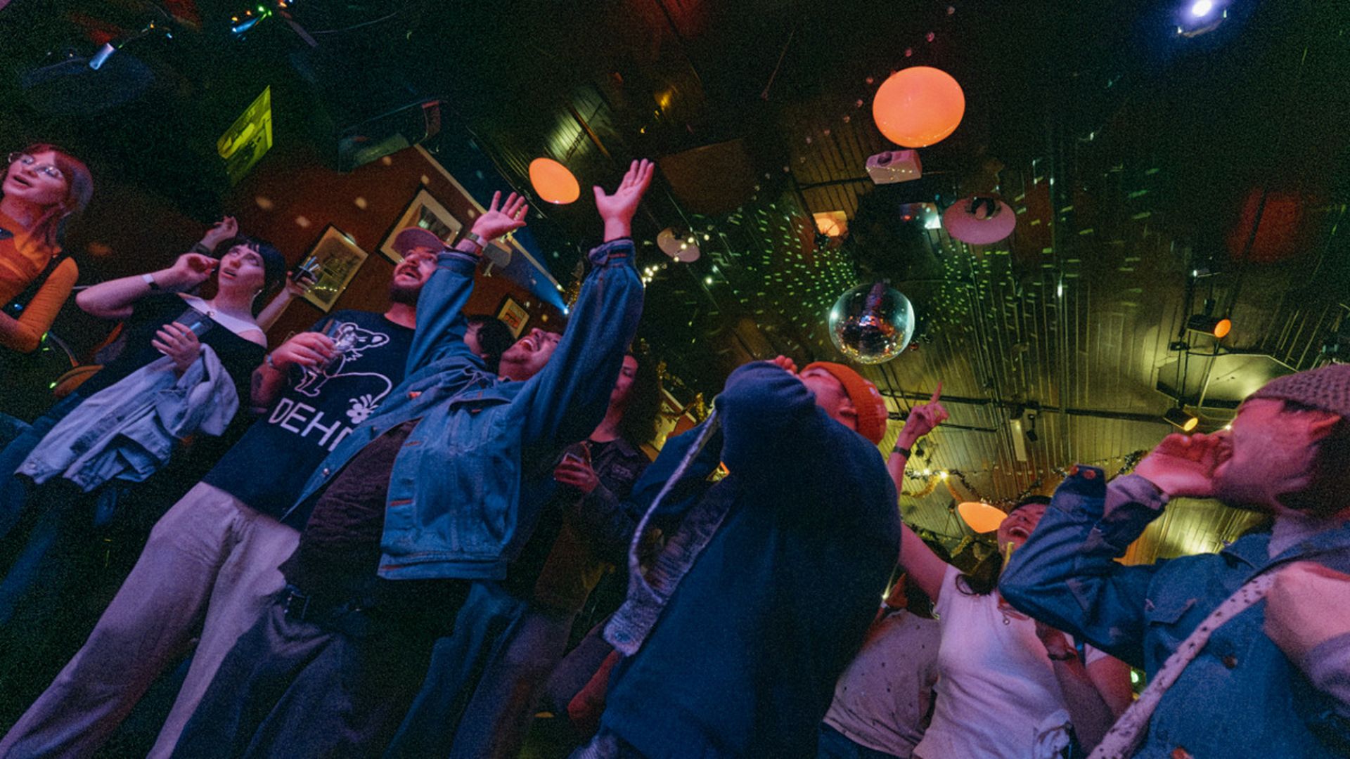 Group of young people enjoying a lively party with colorful disco lights, raised hands, and casual clothing under a ceiling with hanging lamps and a disco ball.