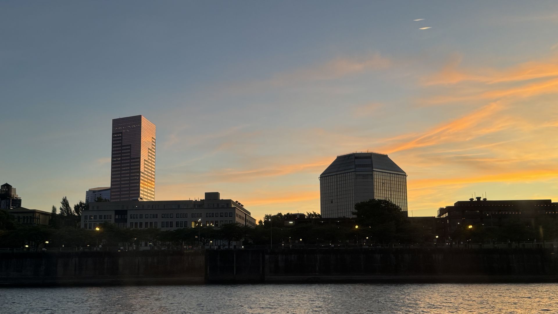 Portland city skyline at sunset with tall buildings, including a reflective rectangular tower and a glass octagonal building, under a sky with orange and blue hues, above calm water.