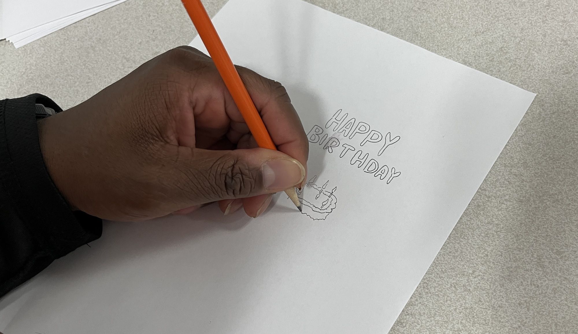 A hand holding an orange pencil is drawing a small birthday cake with candles and the words "HAPPY BIRTHDAY" on a white sheet of paper on a light gray surface.