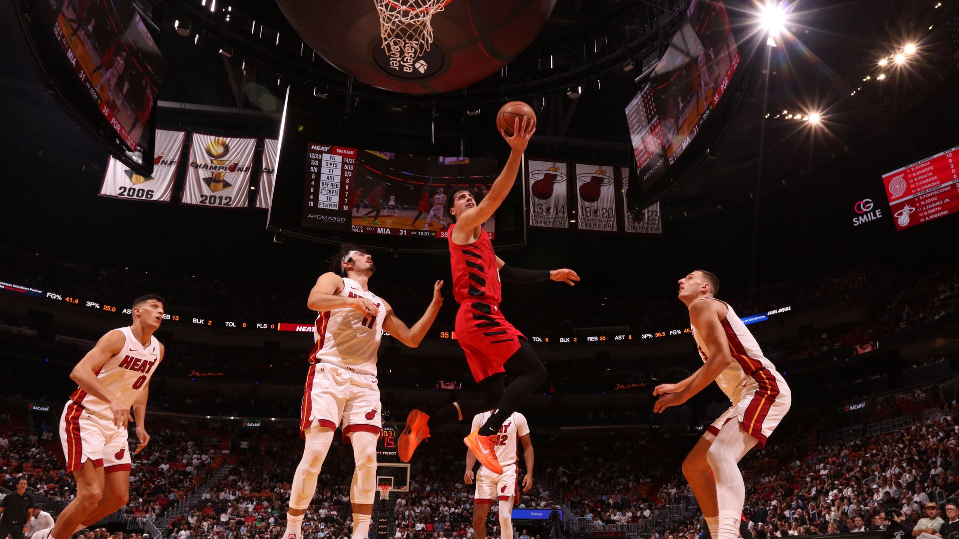 Basketball player in red jumps for a layup near hoop, defended by three players in white Miami Heat jerseys in a crowded arena with championship banners overhead.
