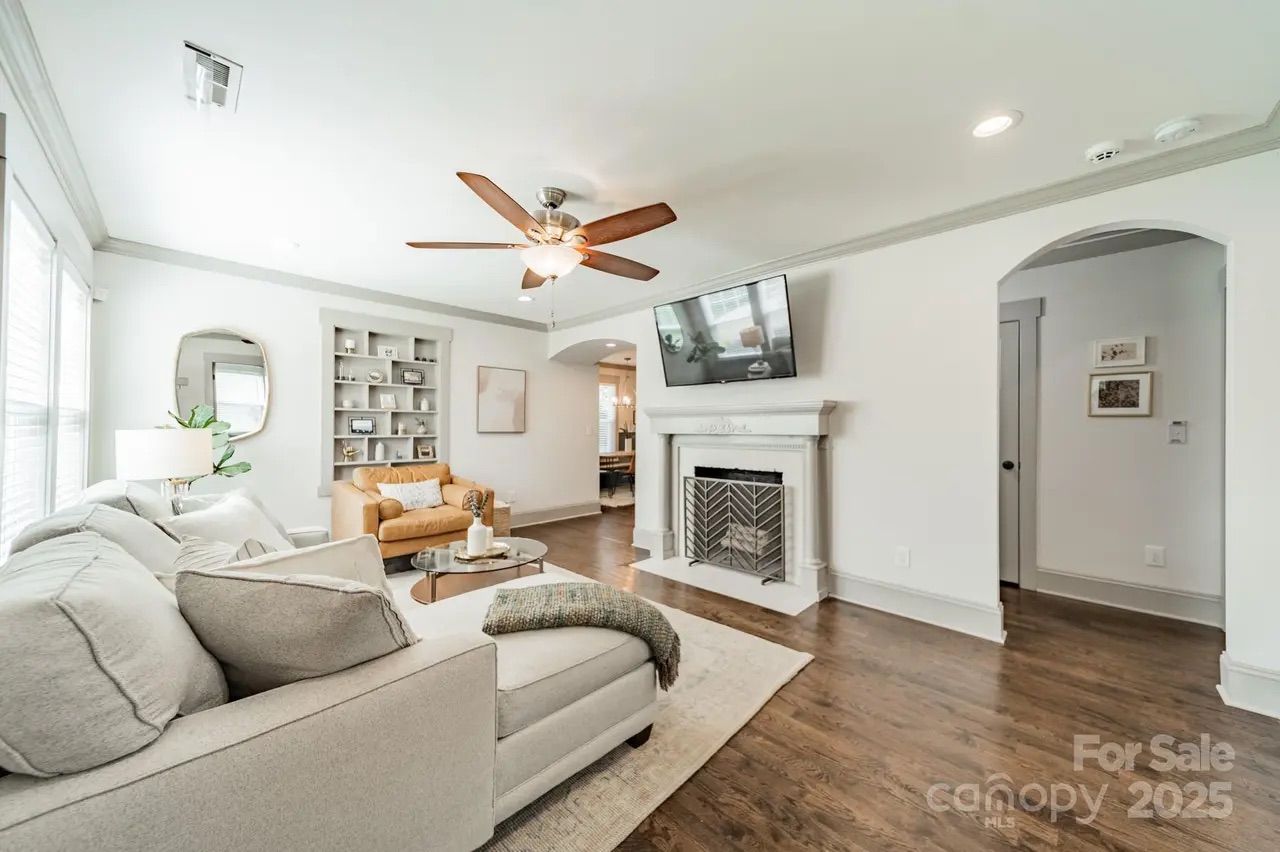 Bright living room with light gray sofa, beige armchair, round glass coffee table, white fireplace, mounted TV, hardwood floor, ceiling fan, and built-in shelves with decor.