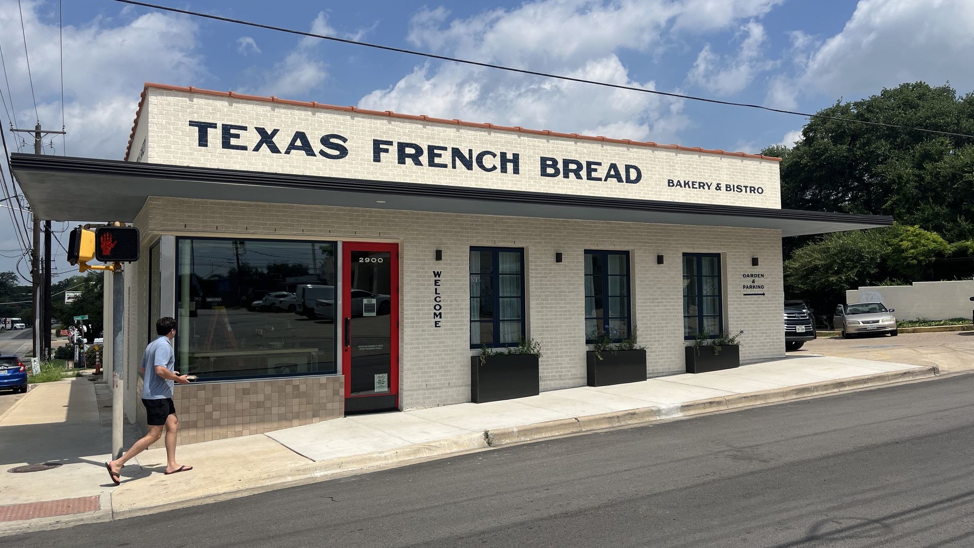 A storefront bakery named "Texas French Bread" with a beige brick facade, red door, and bold black lettering; the sign reads "Bakery & Bistro" as a man walks past planters under a blue sky.