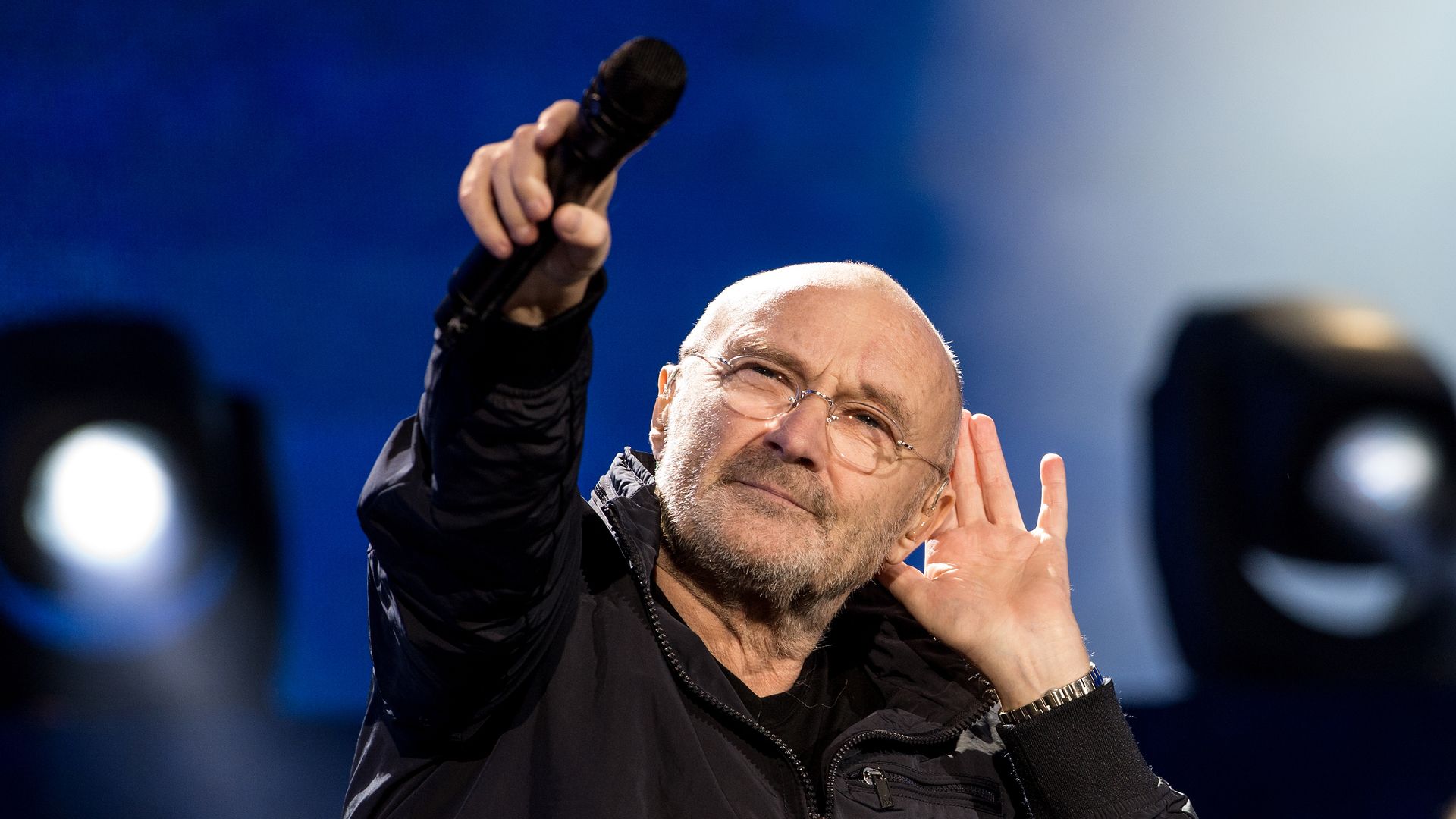 A bald man with glasses and a beard holds a microphone toward the audience and cups his ear, wearing a black jacket on stage with blue and white lights in the background.