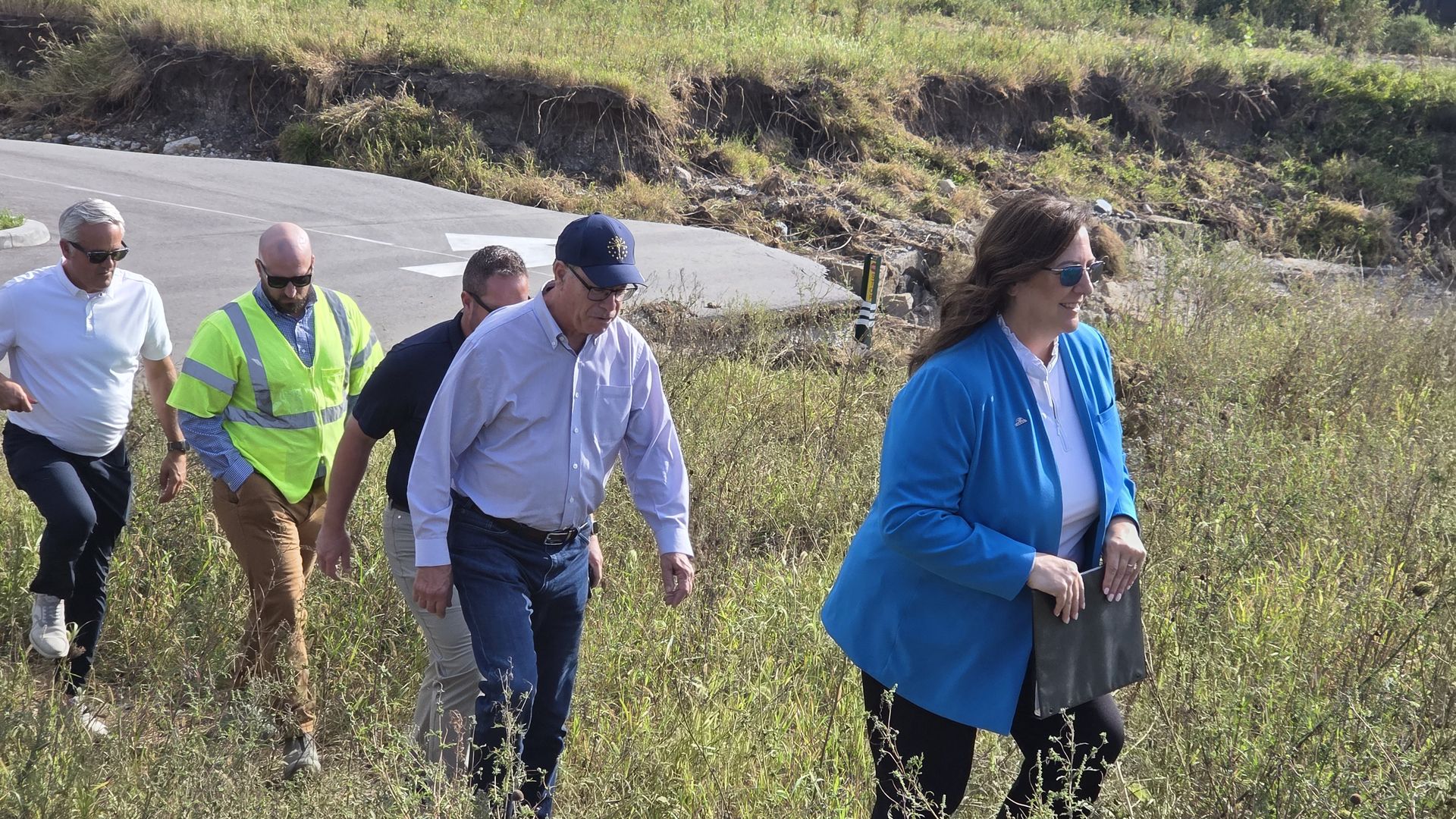 Five adults walk along a rural road bordered by tall grasses; a man in a neon safety vest leads, others in sunglasses wear casual clothes, with a grassy, rocky embankment behind them.