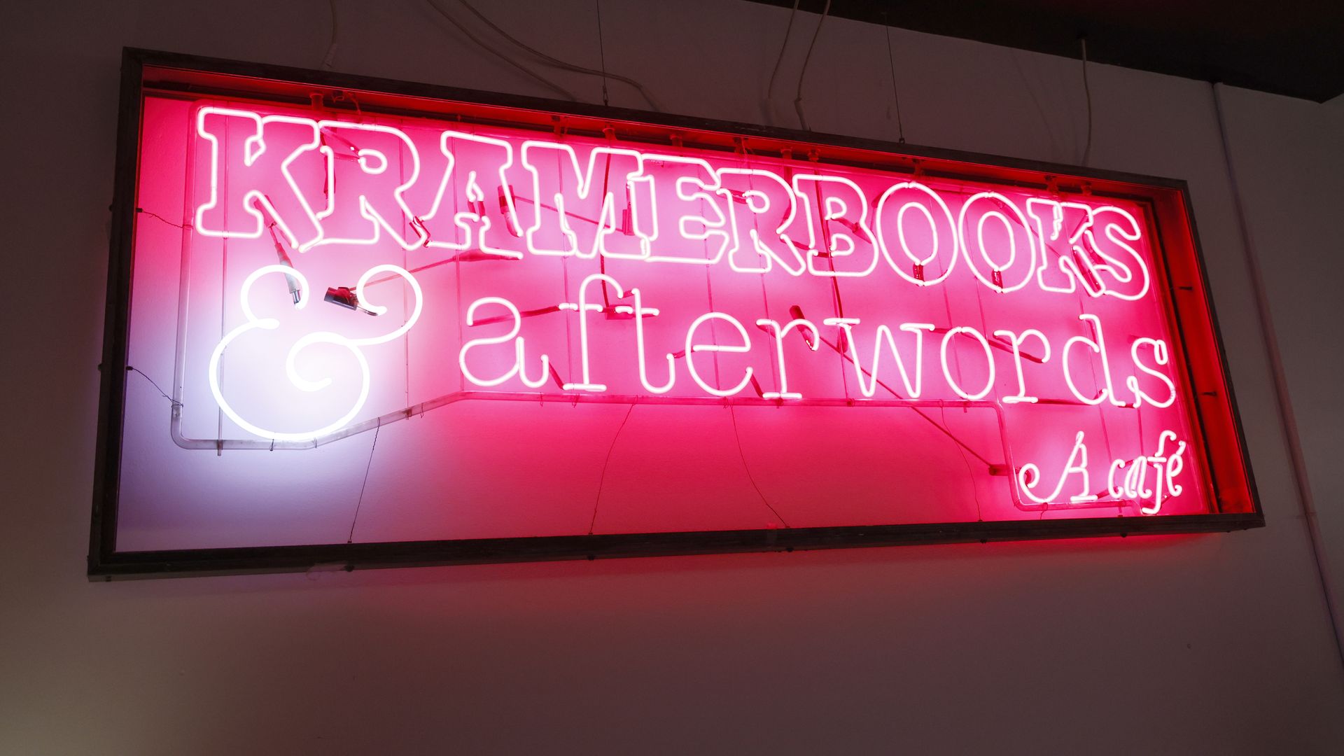A vivid pink neon sign inside a window frame displays bold letters ending with "BOOKS", casting a pink glow on the wall; exposed cables hang from the ceiling in a dim space.