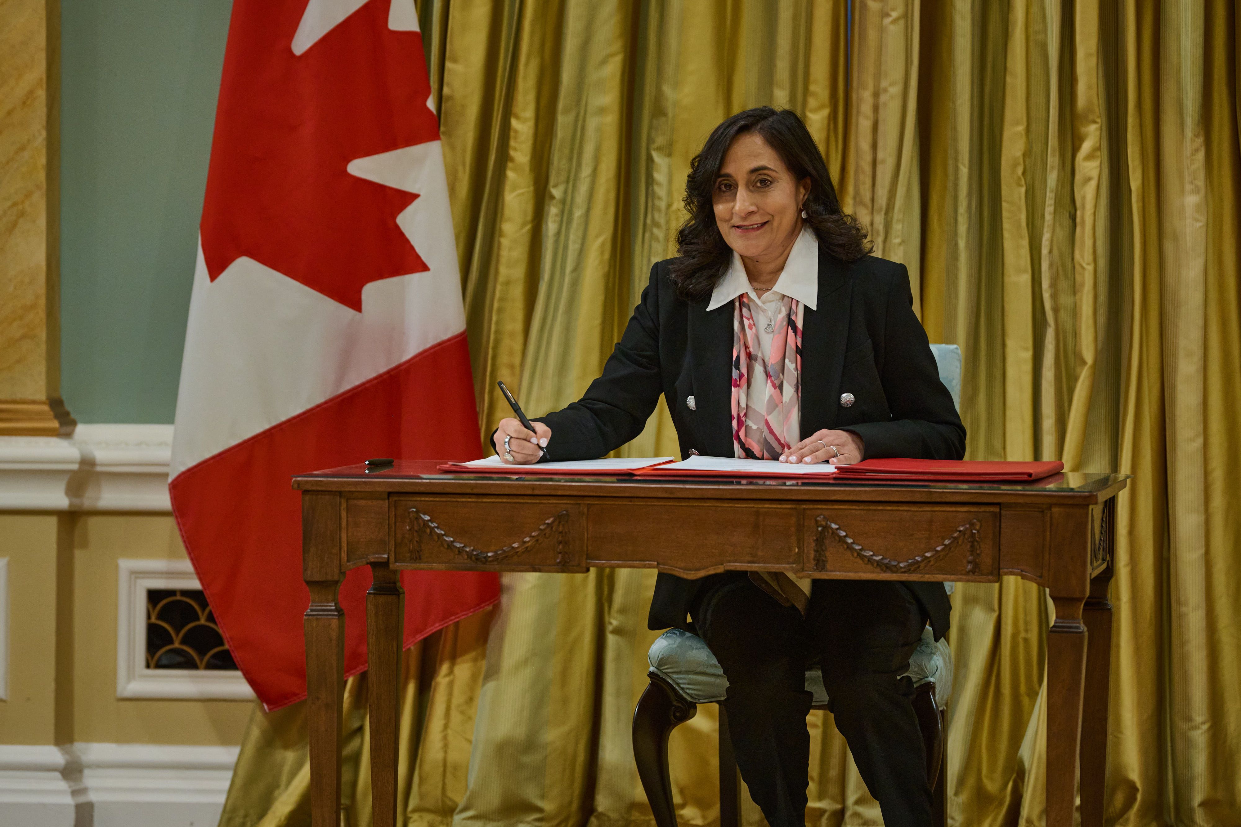 Anita Anand sits at a table with a gold backdrop curtain and a Canadian flag 