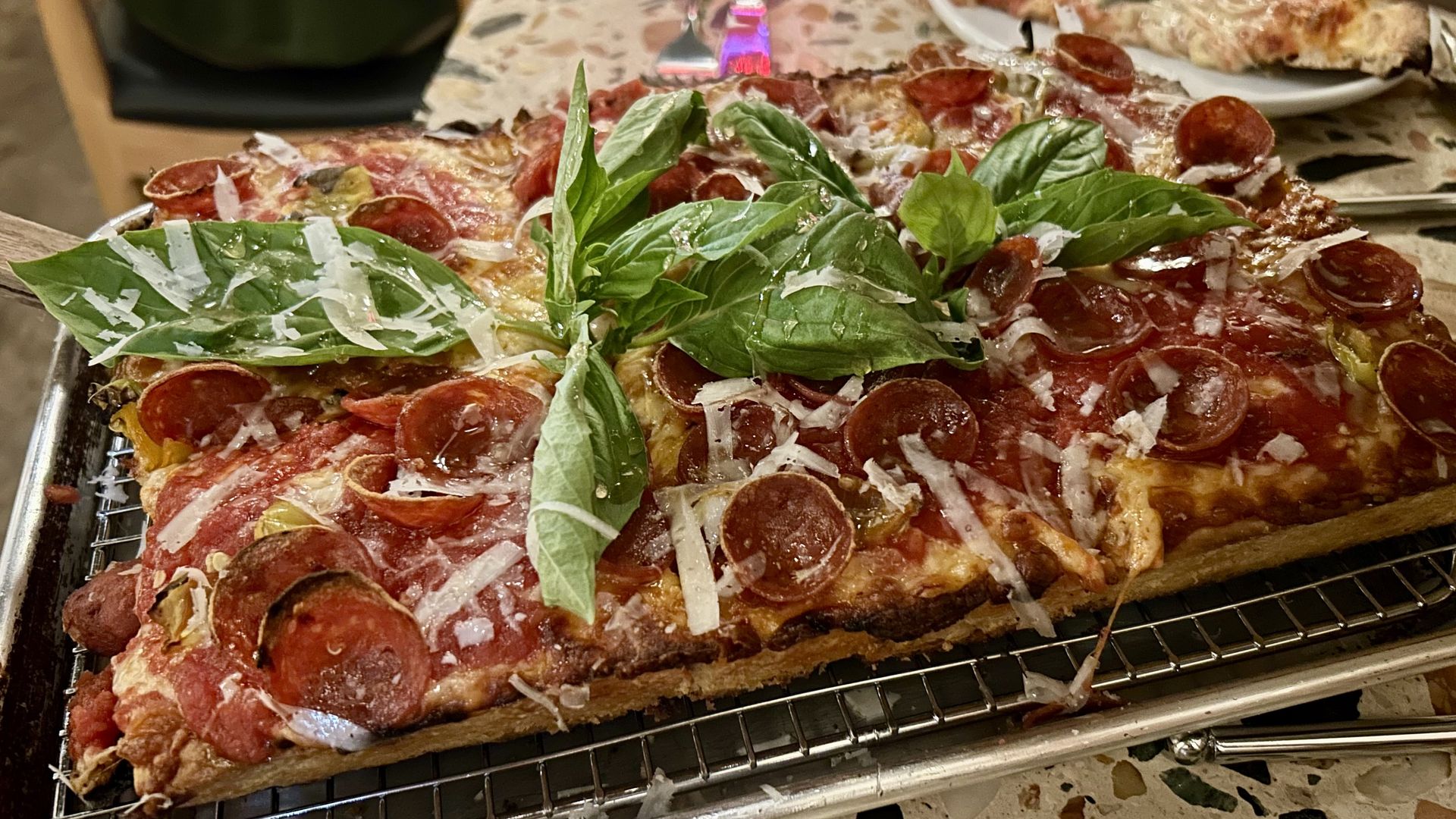 Rectangular pepperoni pizza with melted cheese, tomato sauce, grated cheese, and fresh green basil leaves on a metal rack over a terrazzo table.