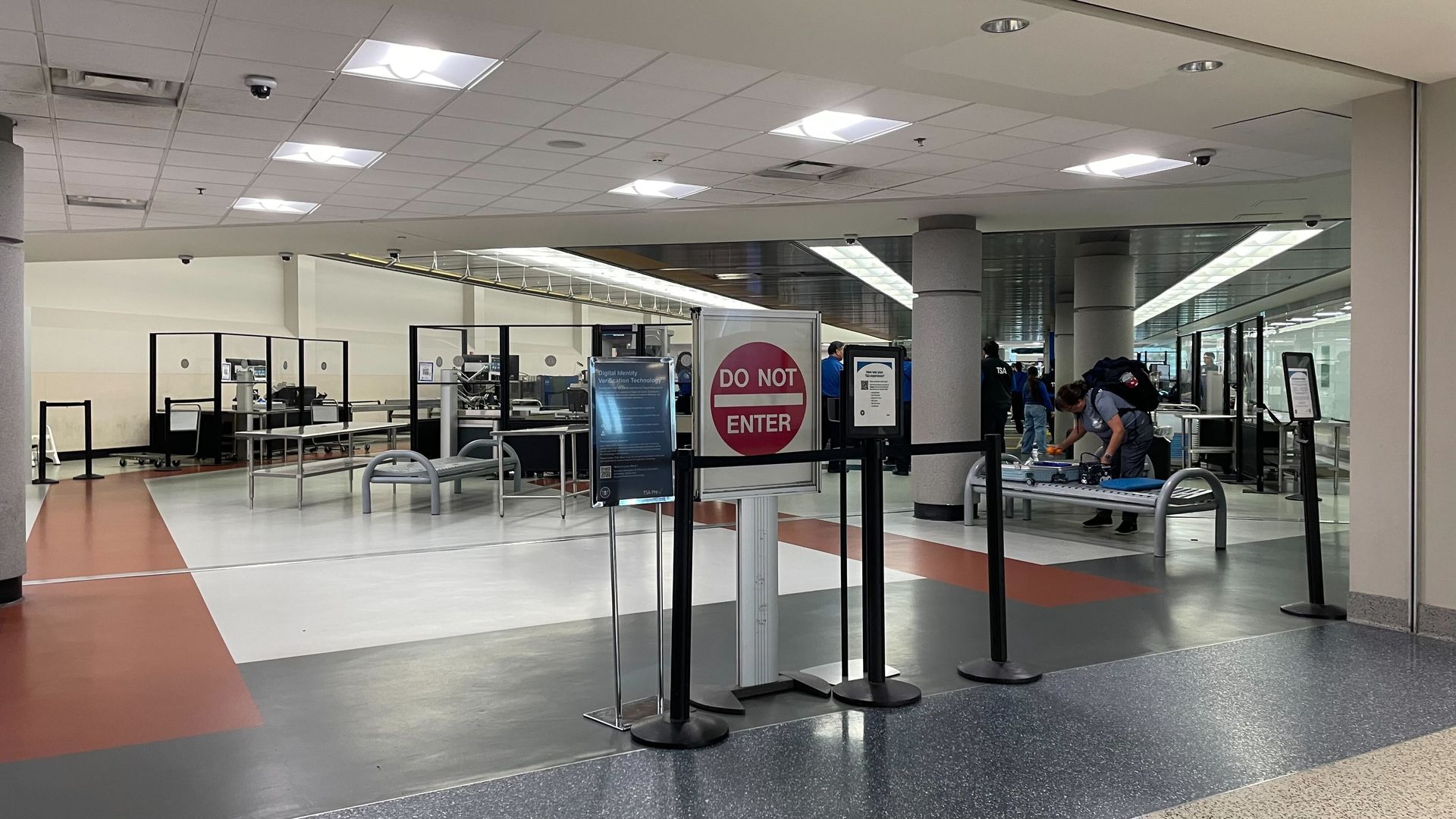 Interior of an airport security checkpoint with a large red DO NOT ENTER sign in the foreground. Black belt barriers, metal desks, and a traveler with a backpack at right; bright ceiling lights.