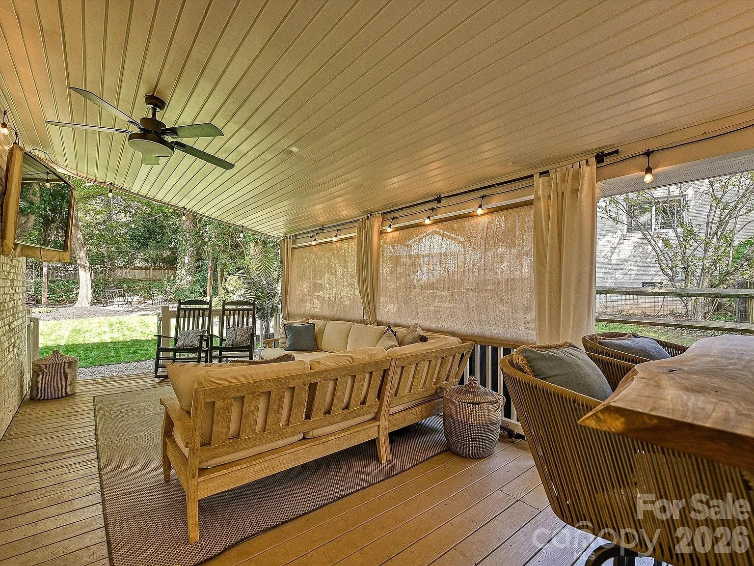 Covered wooden porch with beige seating furniture, two wooden rocking chairs, ceiling fan, string lights, and outdoor view of green lawn and trees.