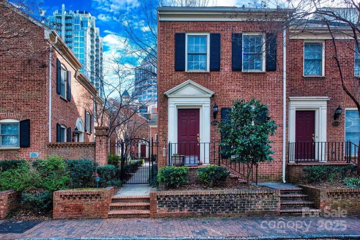 Red brick townhouses with black shutters and burgundy doors, small front steps, green shrubs, leafless trees, and a gated walkway under a blue sky with clouds.