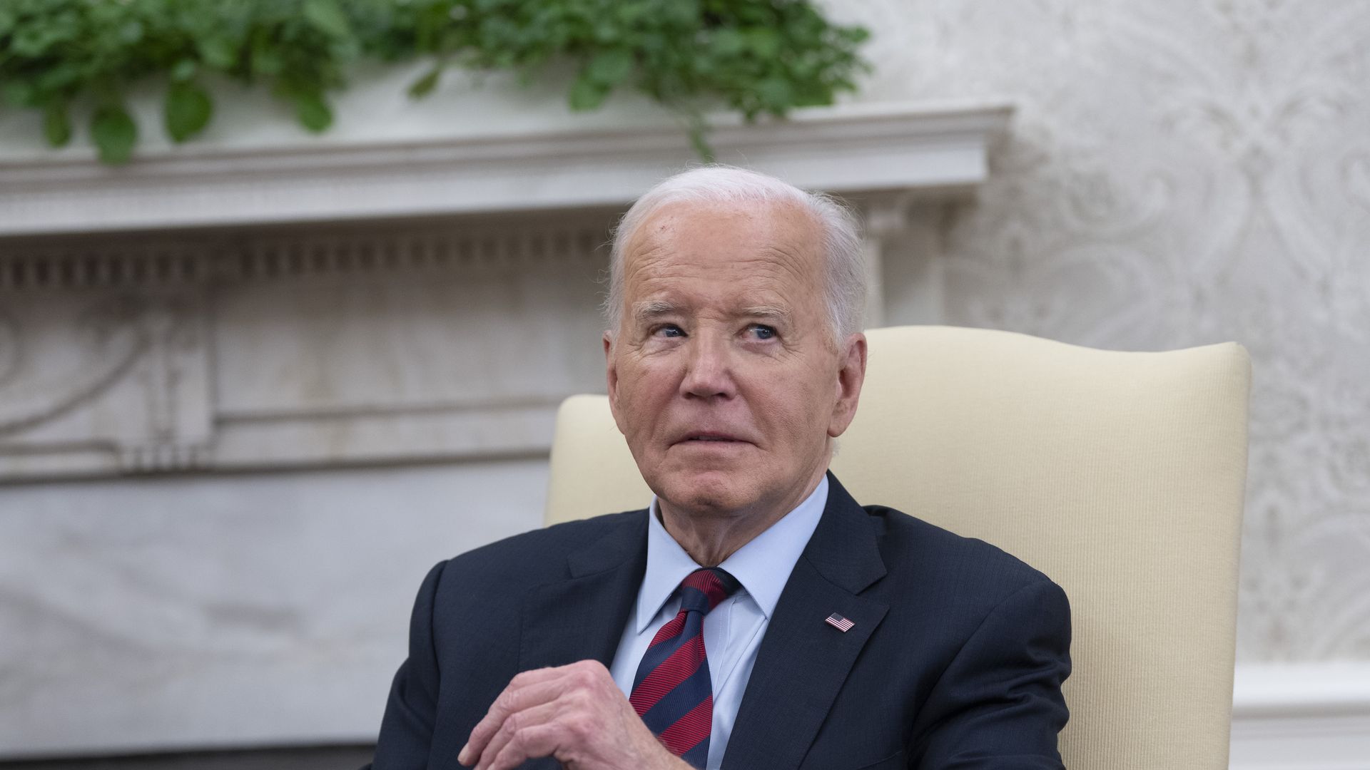 President Joe Biden sits in a beige chair in the Oval office. He has his hand in front of him and is looking off to the side