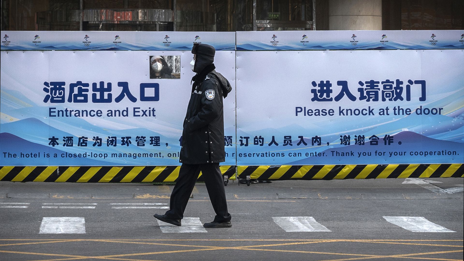 A man looks through a window in a fence as a security guard walks in front of a fenced in hotel ahead of the Winter Olympics. 