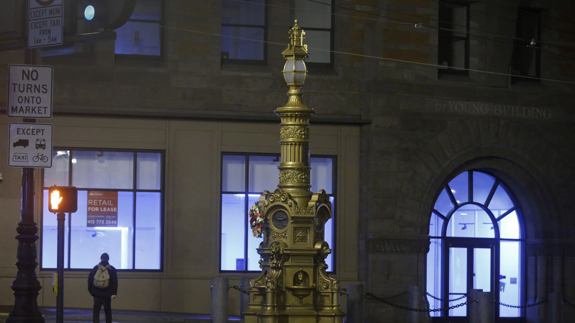 Night street scene featuring a tall ornate gold fountain/lamppost at center, with flowers and chains. Blue-lit storefronts behind; a backpacked pedestrian crosses the street; signs on the left.