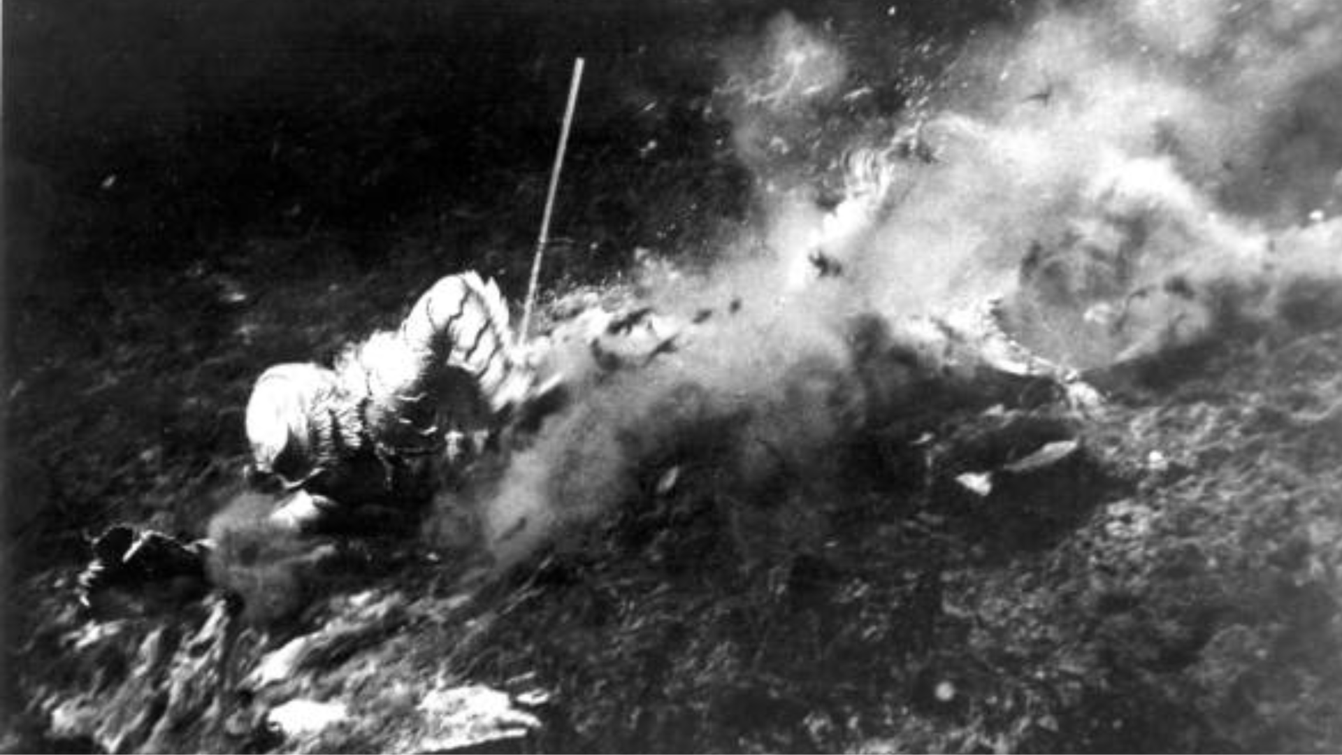 A black-and-white photo of a person in a scaly monster suit thrashing in water, surrounded by bubbles.