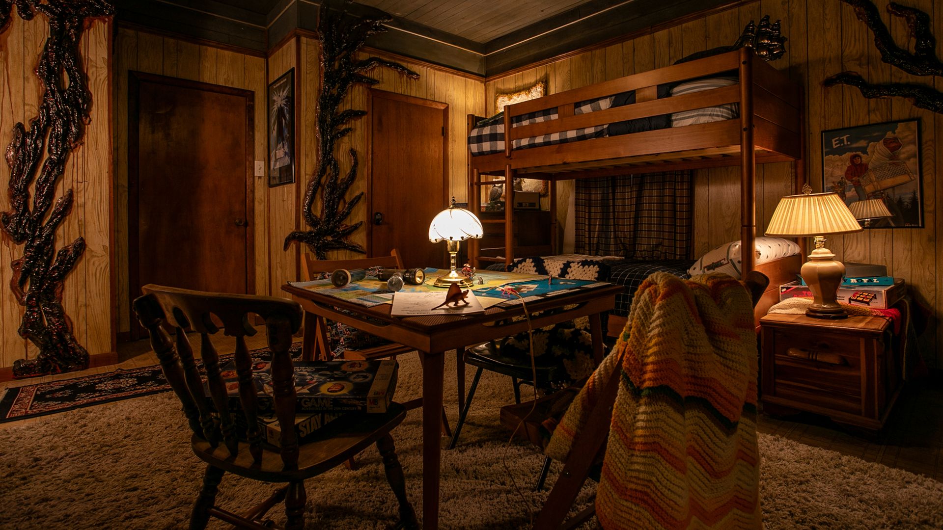 A cozy wood-paneled room with bunk beds, table with board games, vintage lamps, knitted blanket on chair, and eerie black branch decorations on walls under warm lighting.