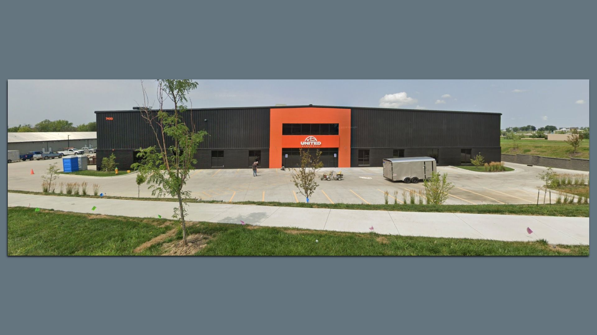 Black and orange commercial building with "United" logo, empty parking lot, a tree in foreground, and a concrete sidewalk under a partly cloudy sky.