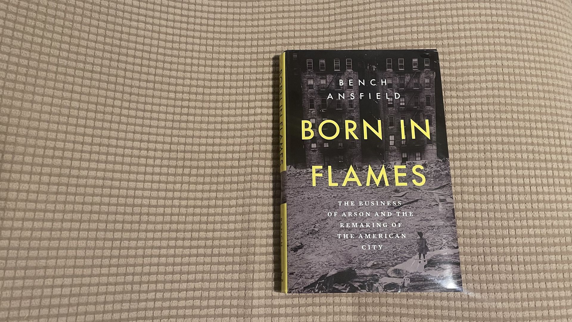 Book cover with a black-and-white city scene and bold yellow lettering reading "BORN IN FLAMES"; beige textured couch in the background, yellow spine labeled "BORN IN FLAMES".