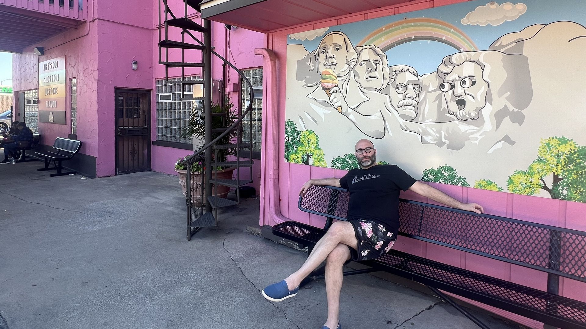 Photo of a man sitting in front of a mural with mountains and a rainbow. 
