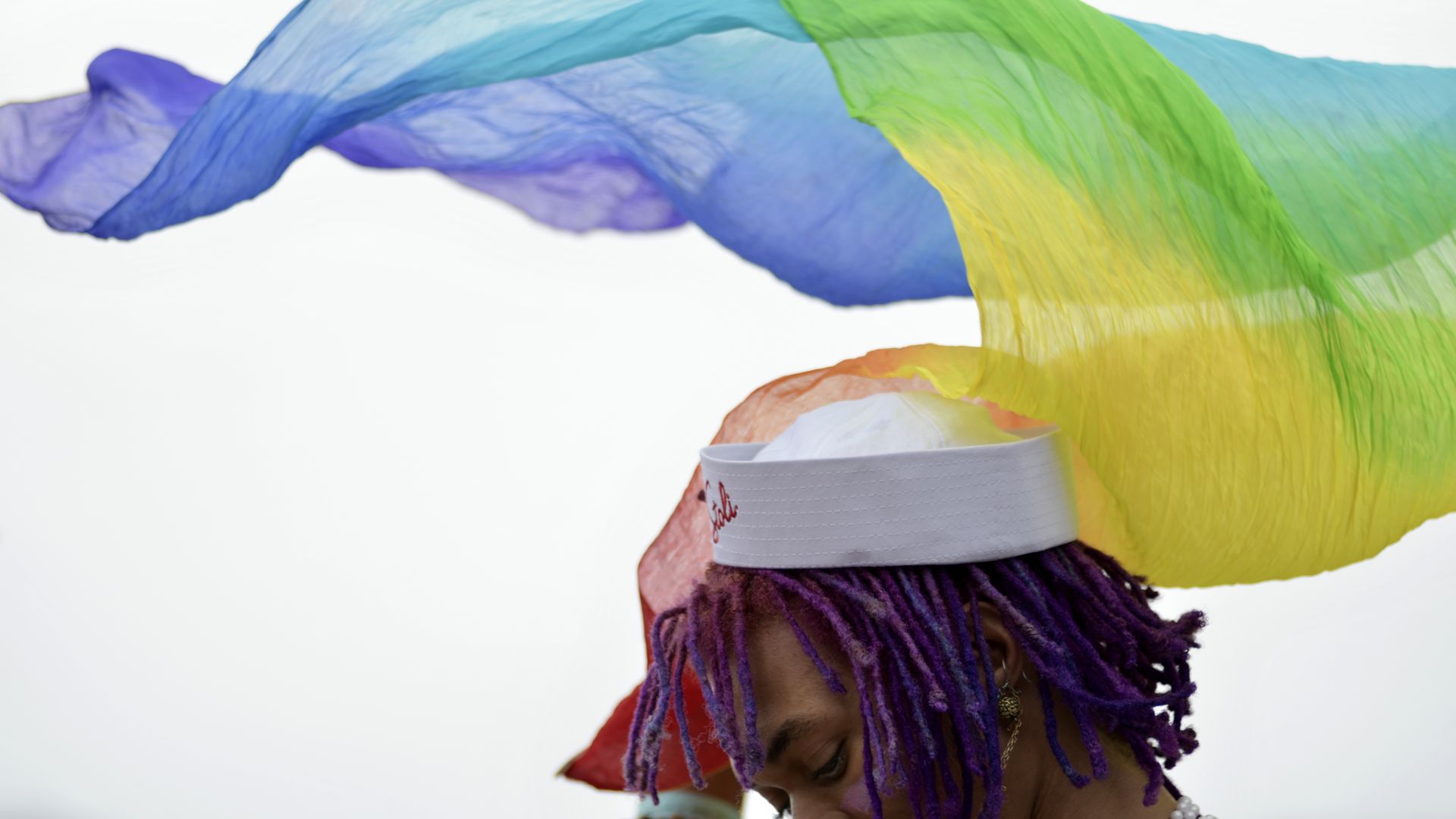 Person with purple braids wearing a white cap; bright sheer ribbons in blue, green, yellow, and orange flow overhead against a pale background.