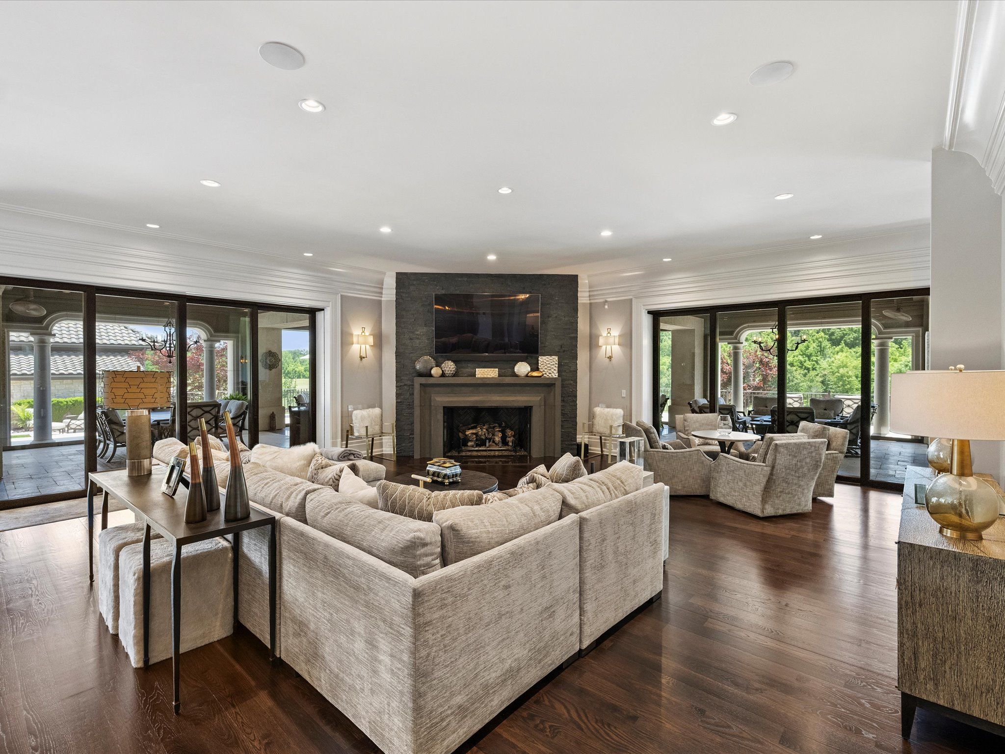 Spacious living room with beige sectional sofa, dark stone fireplace, TV above it, glass sliding doors leading to outdoor patio, wooden floors, and neutral-toned furniture and decor.