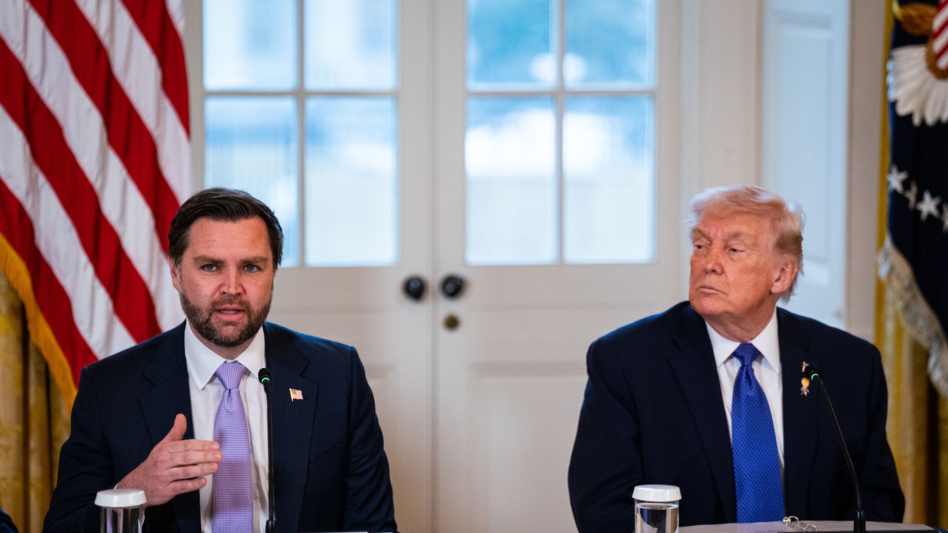 Vance speaking into a microphone while sitting down, wearing a navy suit jacket, white shirt and purple tie, as Trump sits to his right, wearing a navy suit jacket, white shirt and blue tie