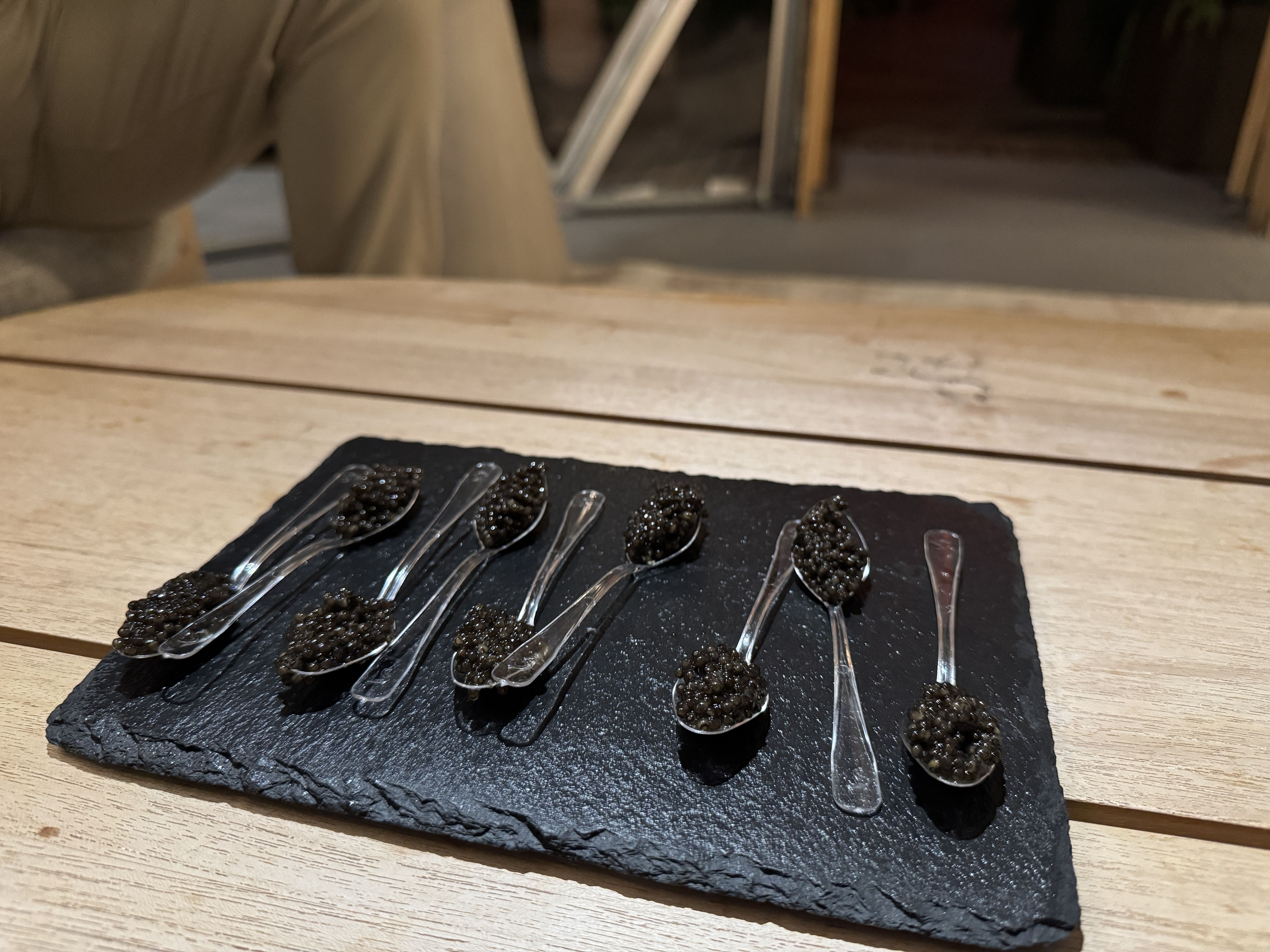 Eight small plastic spoons with black caviar on a black rectangular slate serving tray, placed on a light wooden table with a blurred background.