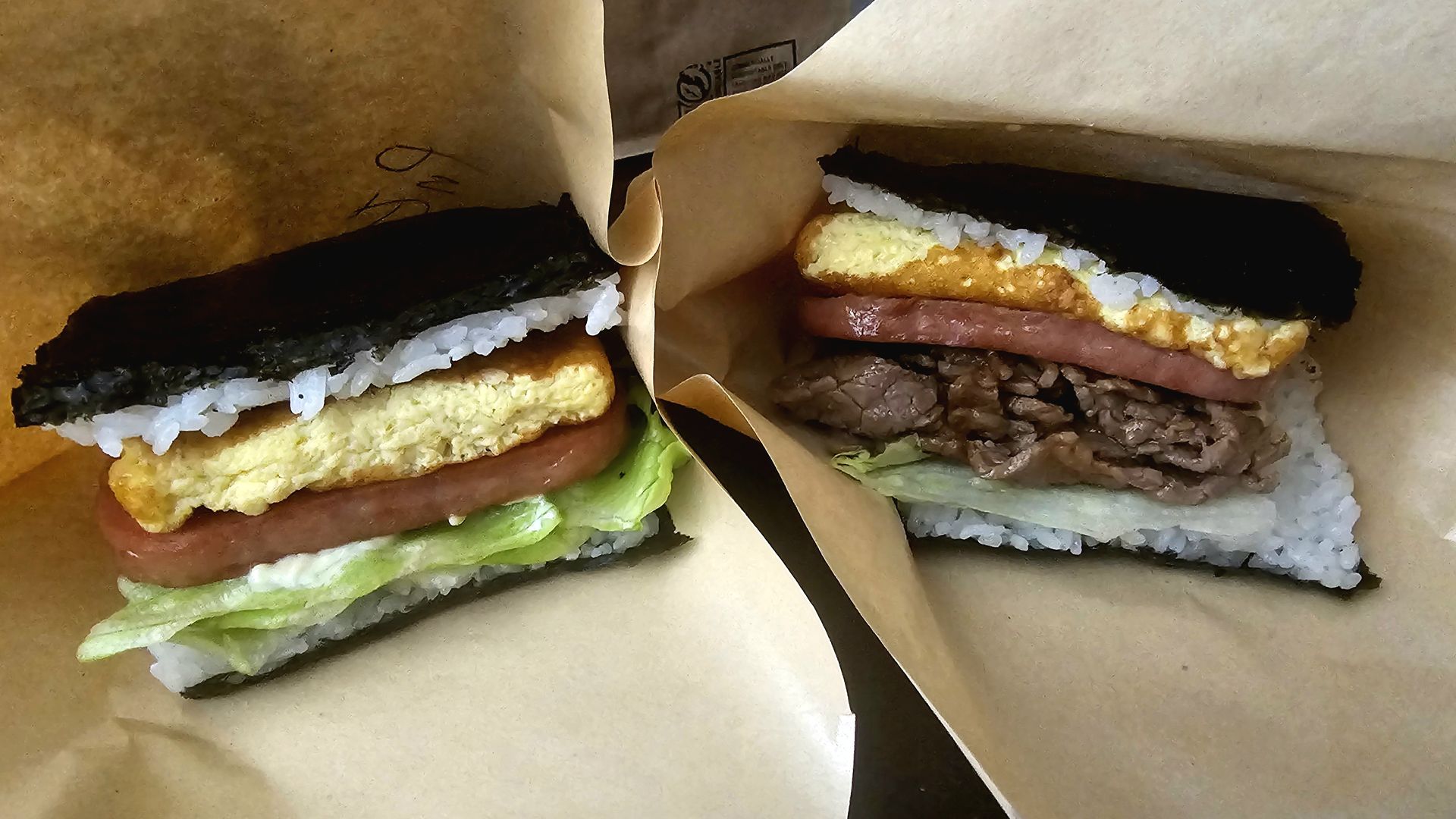 Two rice sandwiches wrapped in brown paper, each with a dark seaweed top, white rice, yellow egg omelette, pink ham, and lettuce visible between layers.