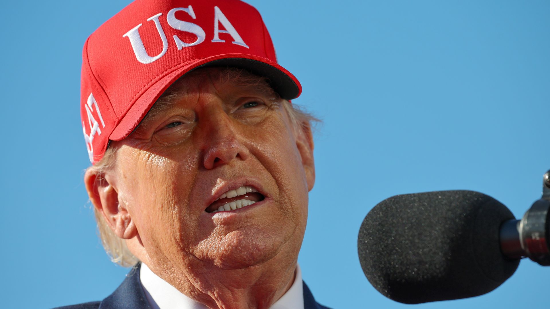 Close-up of President Trump wearing a red cap embroidered with "USA", speaking into a large black microphone, against a clear blue sky; wearing a dark suit and red tie.