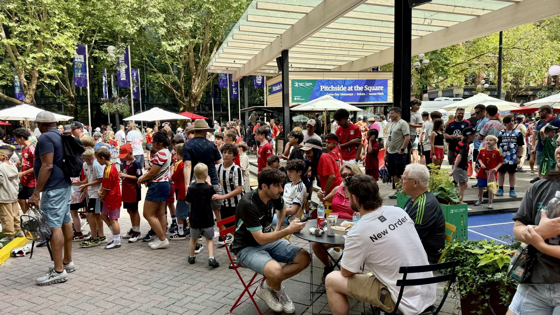 An outdoor crowd of mostly kids and adults in soccer jerseys gathers under a glass canopy, with blue banners reading Pitchside at the Square, tables, umbrellas, and trees in the background.