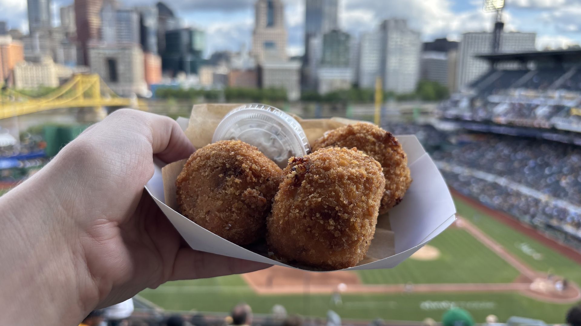 Hand holding a tray with three golden-brown fried snacks and a small container of white dipping sauce, overlooking a baseball stadium with a yellow bridge and city skyline in the background.