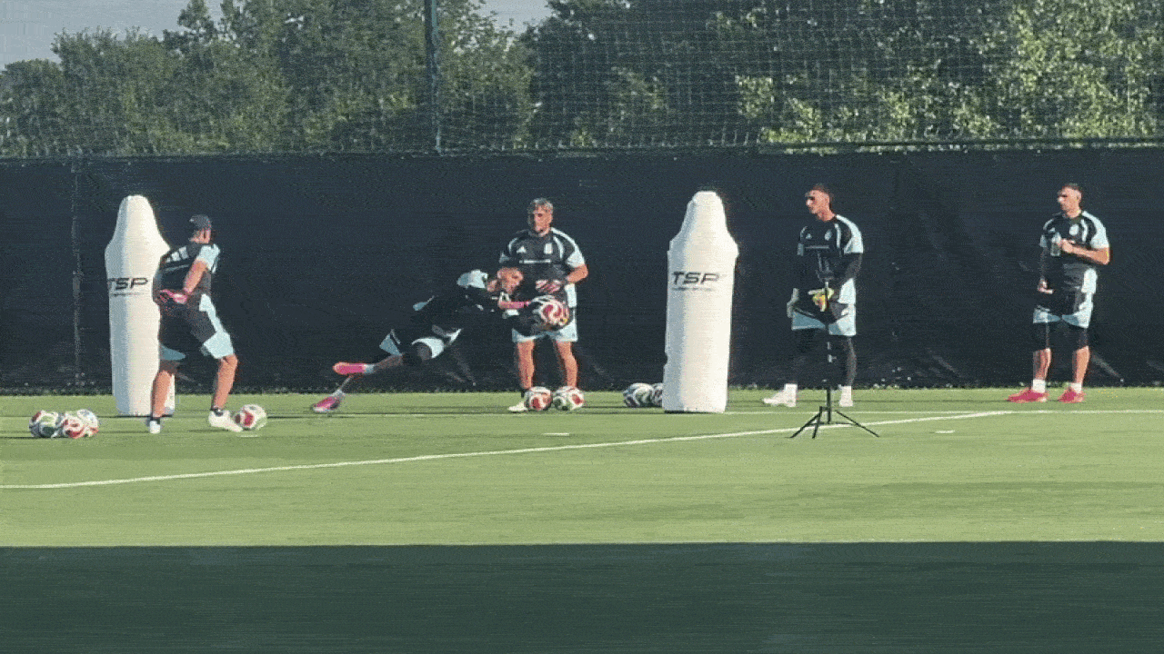Soccer training on a sunny field: a goalkeeper dives to block a ball while teammates watch; white inflatable dummies, scattered balls, and a tripod on the green pitch.