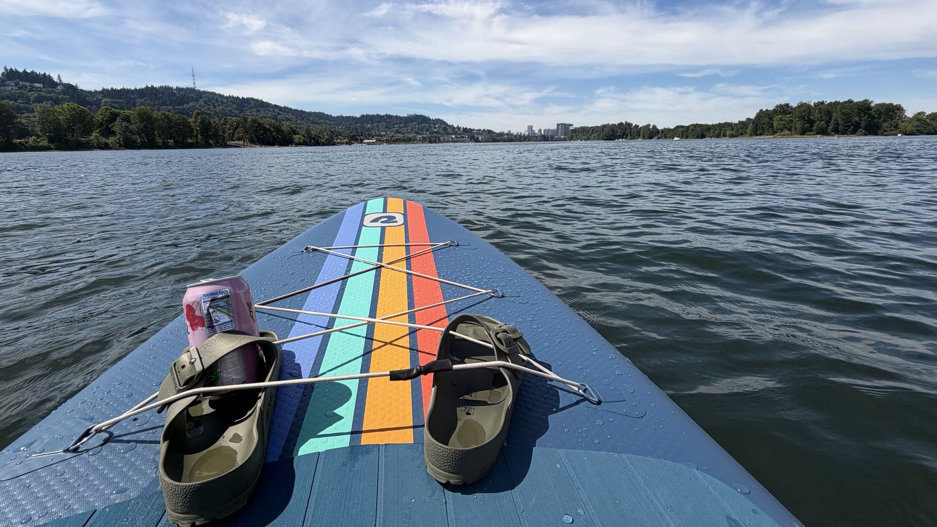 From a paddleboard on a lake: a blue board with bright stripes, sandals and a drink on the deck, calm water, tree-lined shore, distant city skyline, and a clear blue sky with wispy clouds.