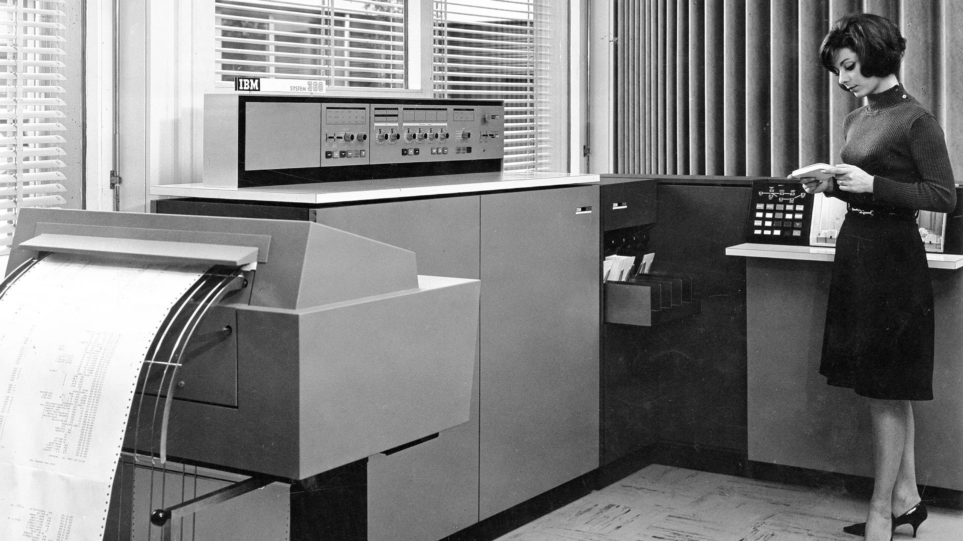 Black-and-white photo of an IBM System/360 mainframe console; a woman in a dark sweater reads a document beside the machine, with a roll-fed printer and blinds in the background.