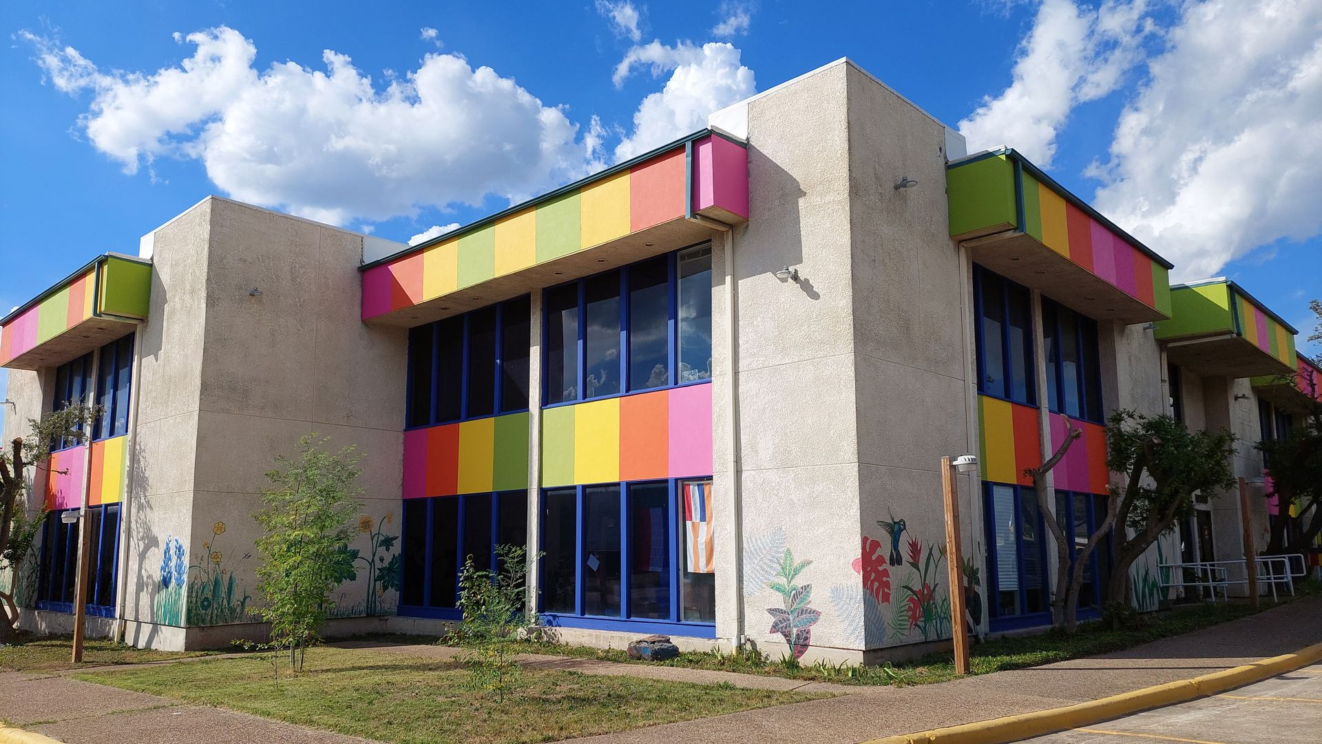 Two-story beige building with colorful rainbow stripe accents and blue window frames under a partly cloudy blue sky. Sidewalk, grass, and small trees surround the building.
