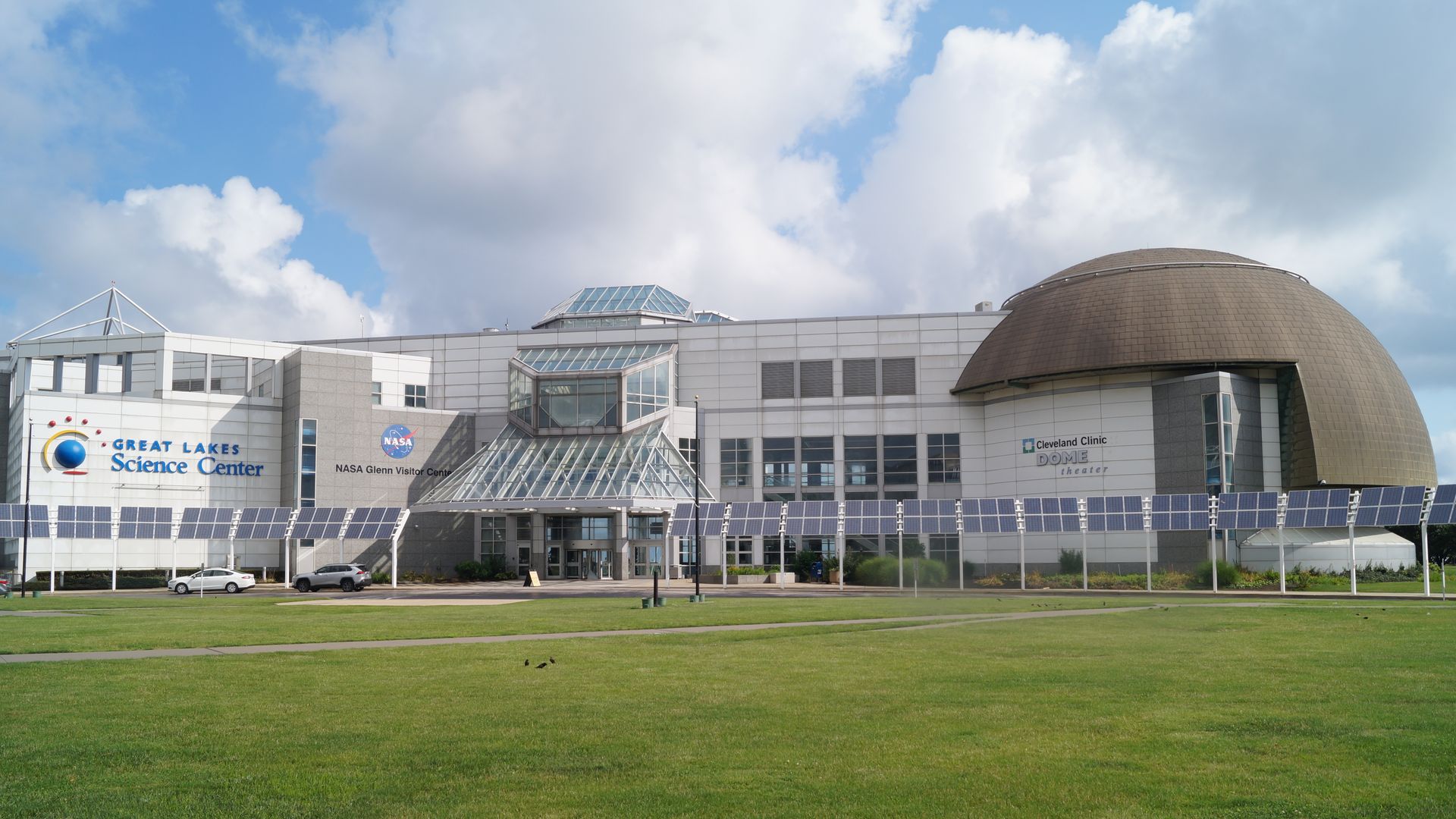 Wide view of the Great Lakes Science Center with solar panels in front and the Cleveland Clinic Dome theater on a partly cloudy day.