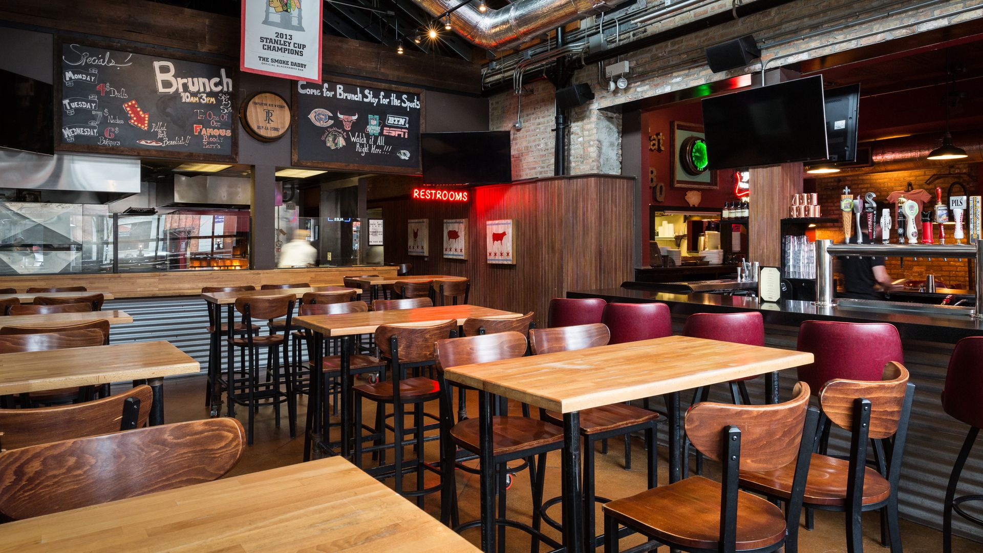Empty sports bar restaurant with wooden tables and chairs, large ductwork, Chicago Blackhawks banner, chalkboard brunch menus, beer taps, and a red "RESTROOMS" sign.