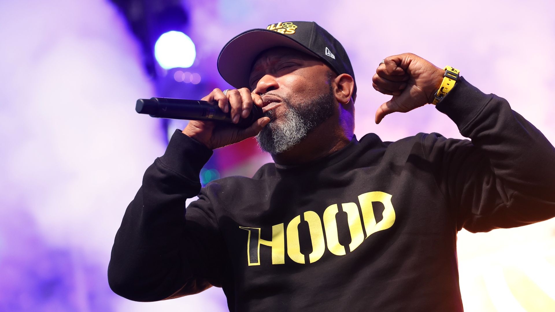 Rapper Bun B, wearing a black longsleeve shirt with yellow lettering that says "HOOD," a yellow watch and "Trill Burgers" black and yellow hat, raps into a microphone with a purple background