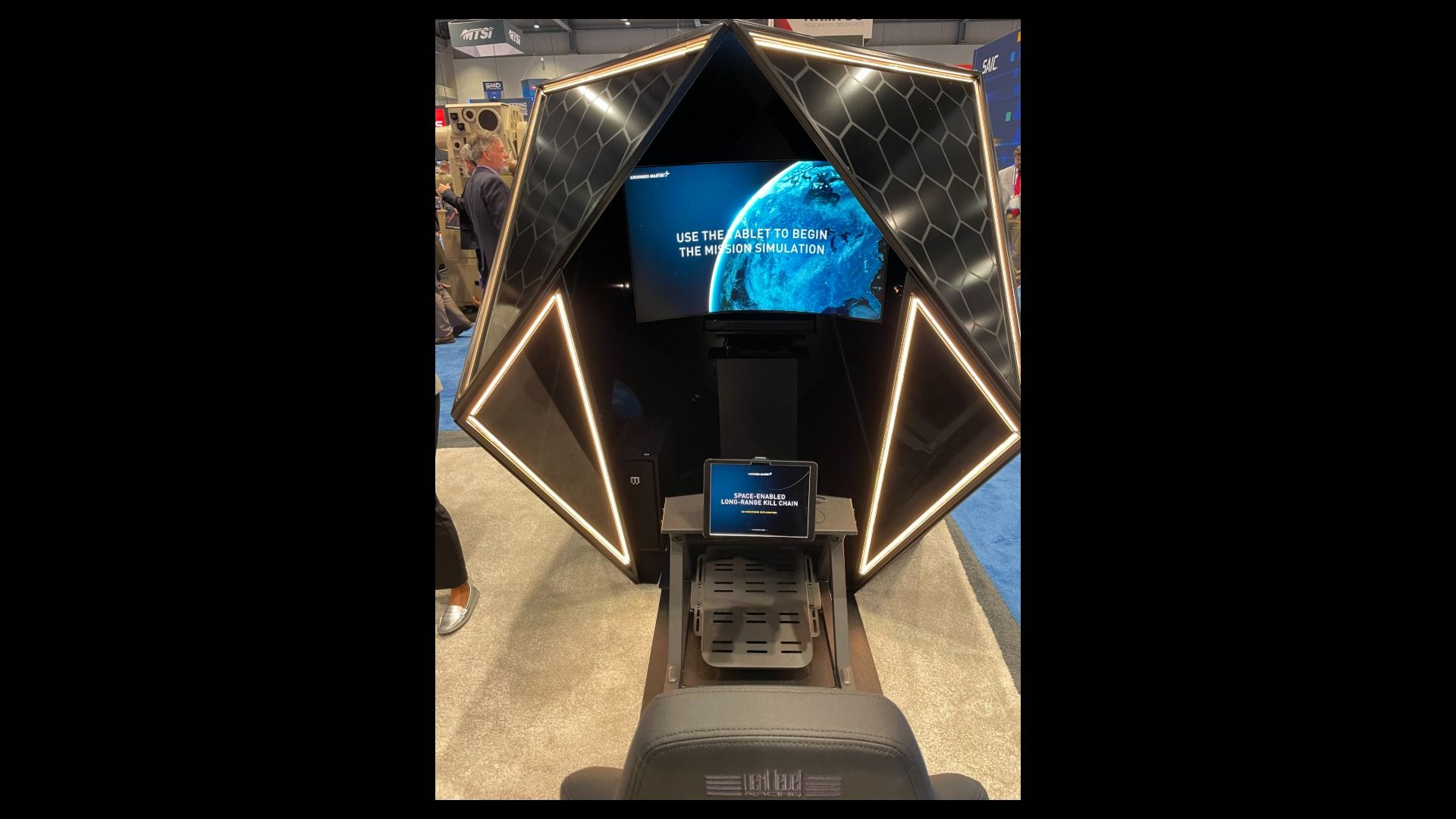 Black geometric booth with honeycomb panels and lit edges. A screen shows Earth and the instruction "Use the tablet to begin the mission simulation." A tablet on a stand sits before a chair.