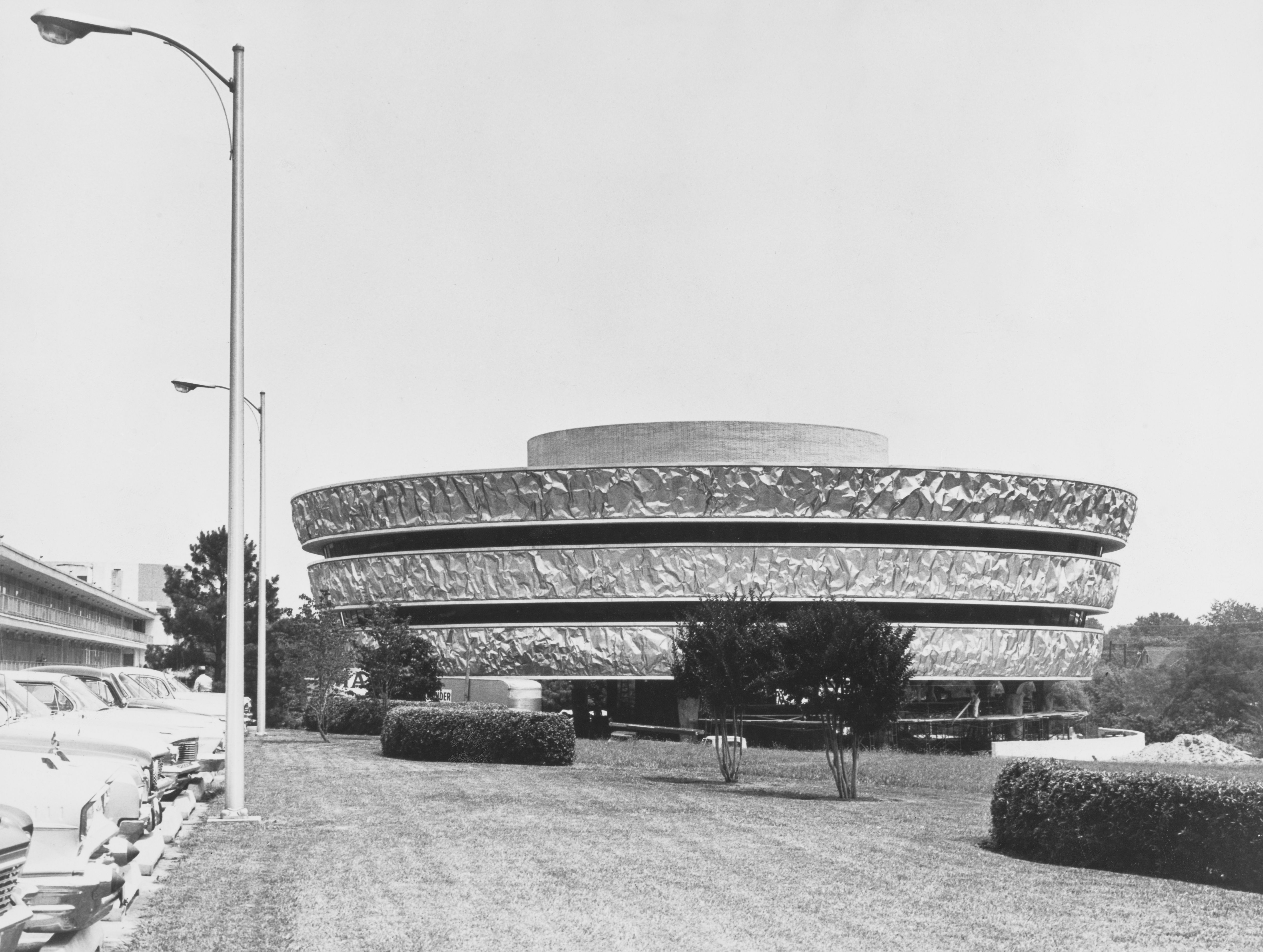 Black-and-white image of a large textured circular, multi-tier fountain in an open area; a tall street lamp stands to the left, with parked cars and trees in the background.